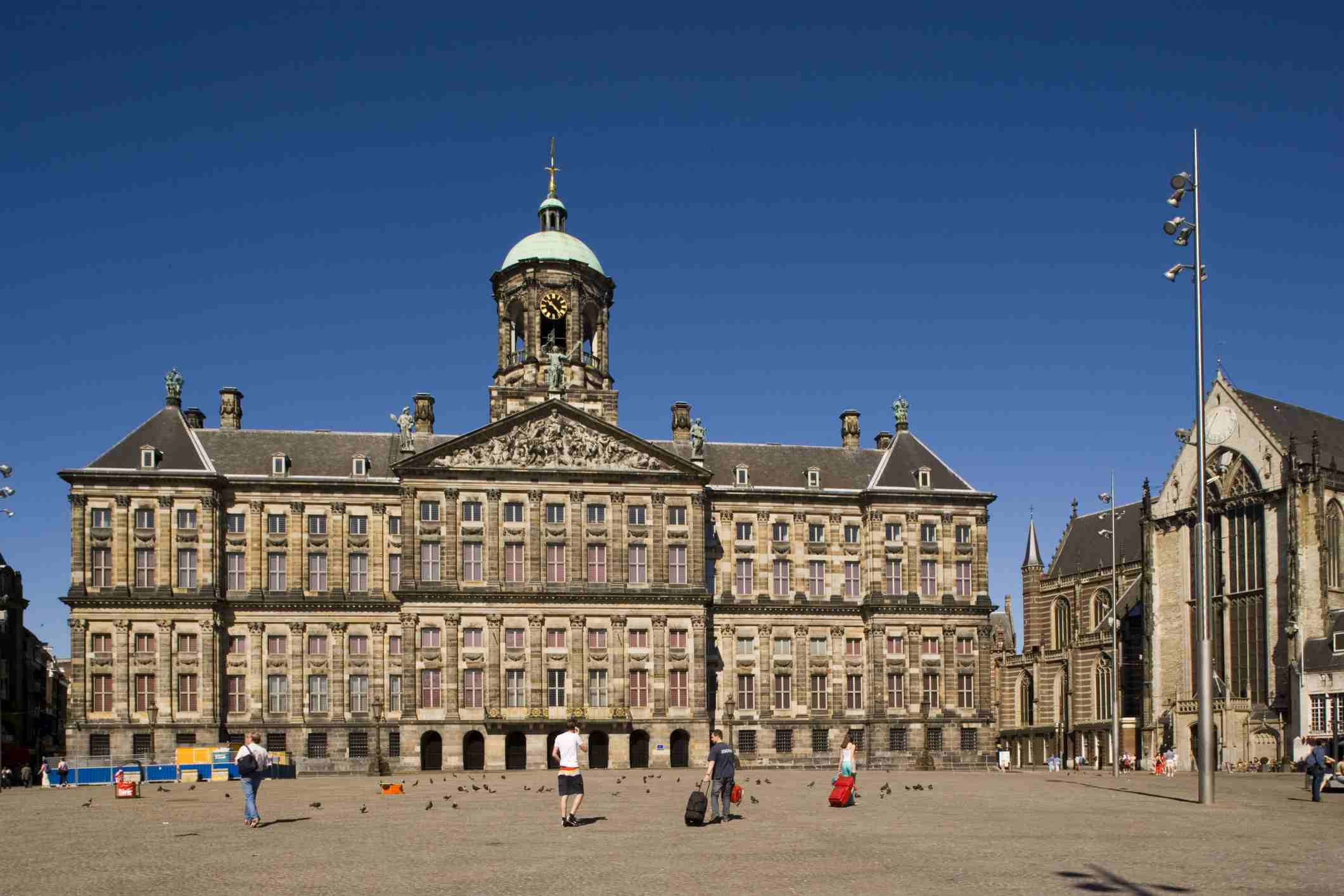 Amsterdam 桌布 壁紙 — 宏偉的歷史宮殿，擁有華麗的外牆和綠色圓頂，矗立在清澈的藍天下，人們在鋪砌的廣場上與鴿子漫步。