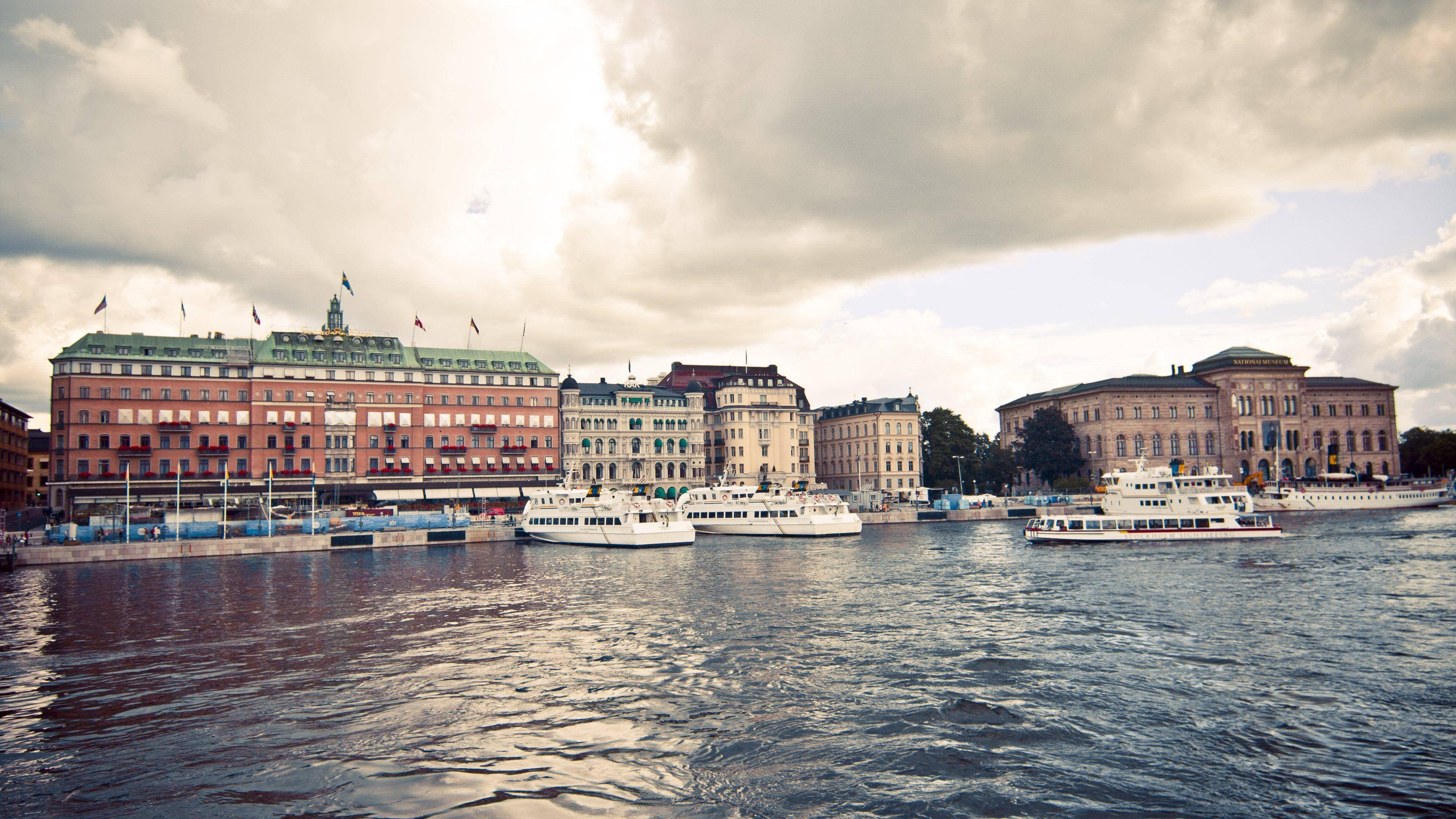 Sweden 桌布 壁紙 — 宏偉的歷史建築沿著熱鬧的海濱排列，白色船隻在波光粼粼的水面上滑行，天空佈滿戲劇性的雲層。