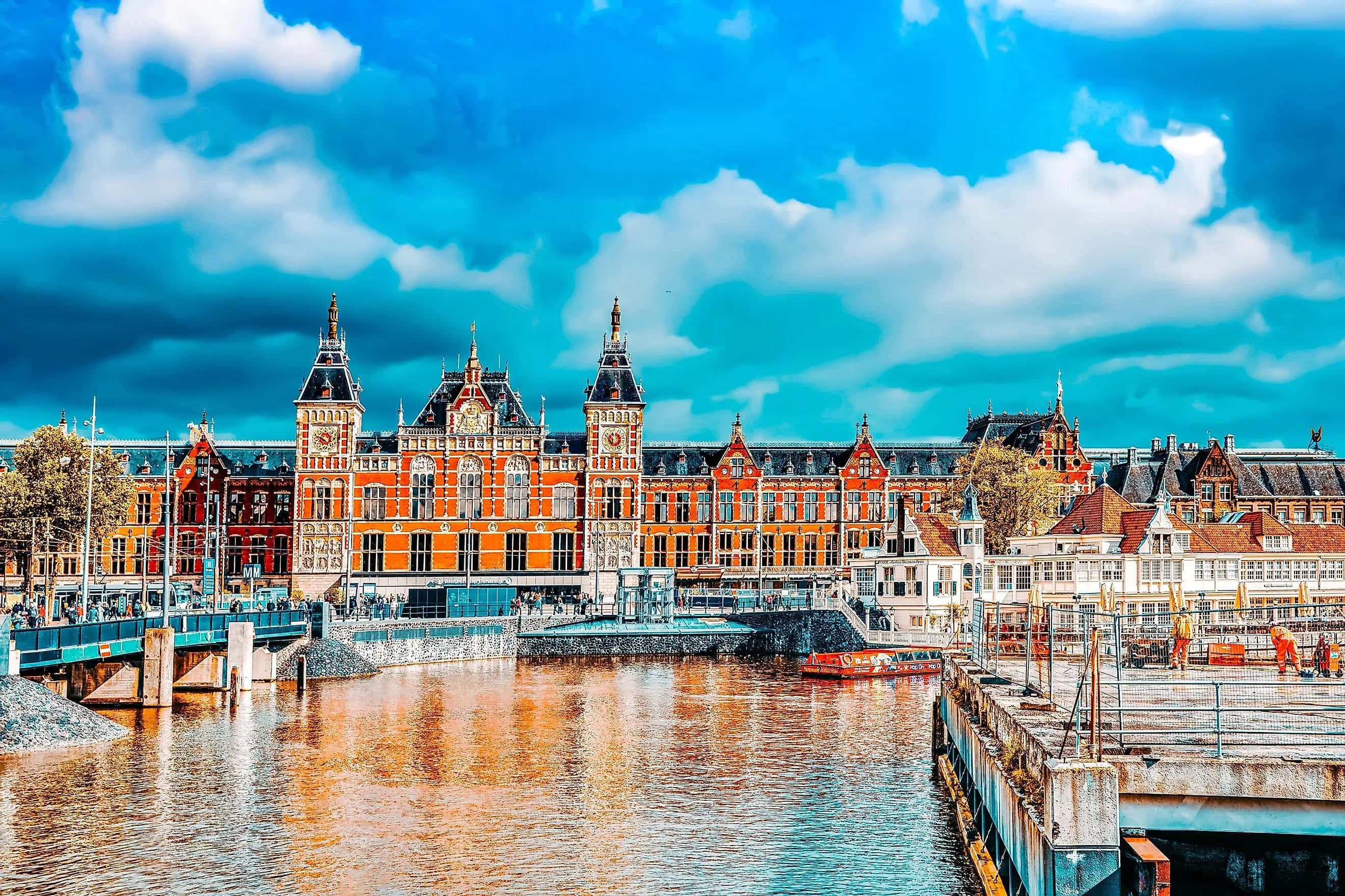 Netherlands 桌布 壁紙 — 宏偉的歷史火車站，紅磚塔樓雄偉地矗立在寧靜的運河旁，上方是湛藍的天空和蓬鬆的白雲。