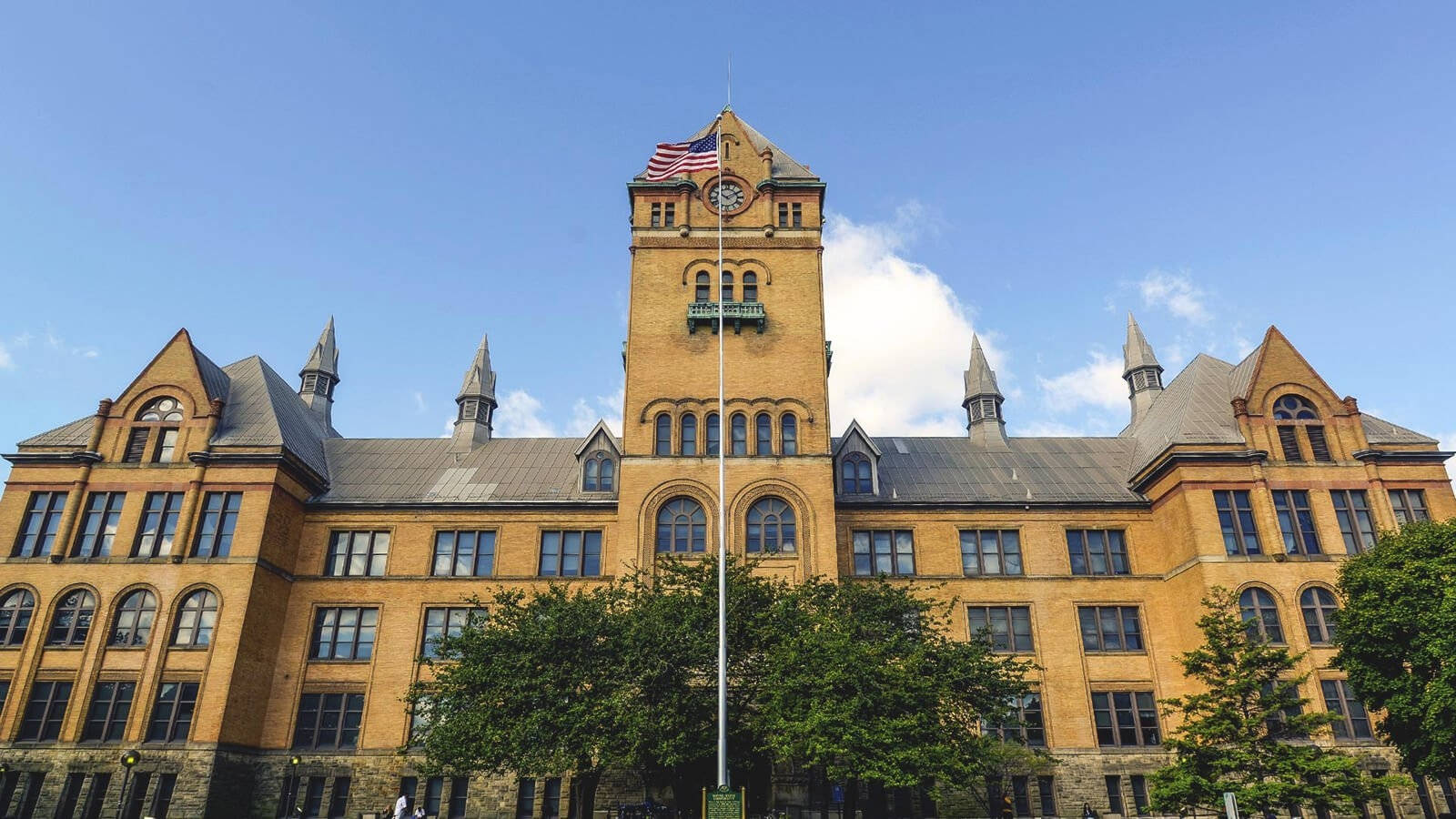建築 桌布 壁紙 — 宏偉的歷史磚造大學建築，設有鐘樓和美國國旗，在晴朗的藍天下。