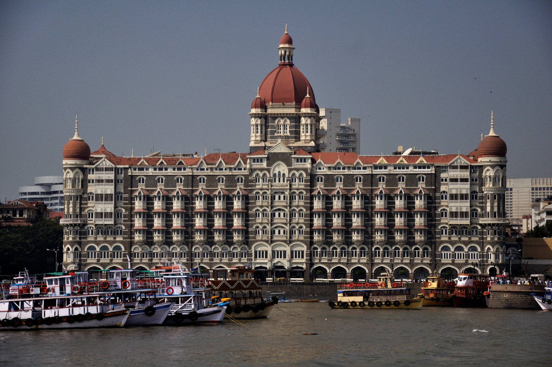 Mumbai 桌布 壁紙 — 宏偉的殖民風格泰姬陵皇宮酒店，紅色圓頂，在朦朧的天空下，雄偉地矗立在一個充滿色彩繽紛船隻的港口旁。