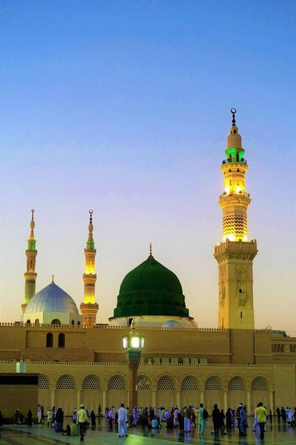 Madina 桌布 壁紙 — 宏偉的清真寺，擁有綠色和藍色的圓頂和發光的宣禮塔，在暮色天空下閃耀，信徒在下方聚集。