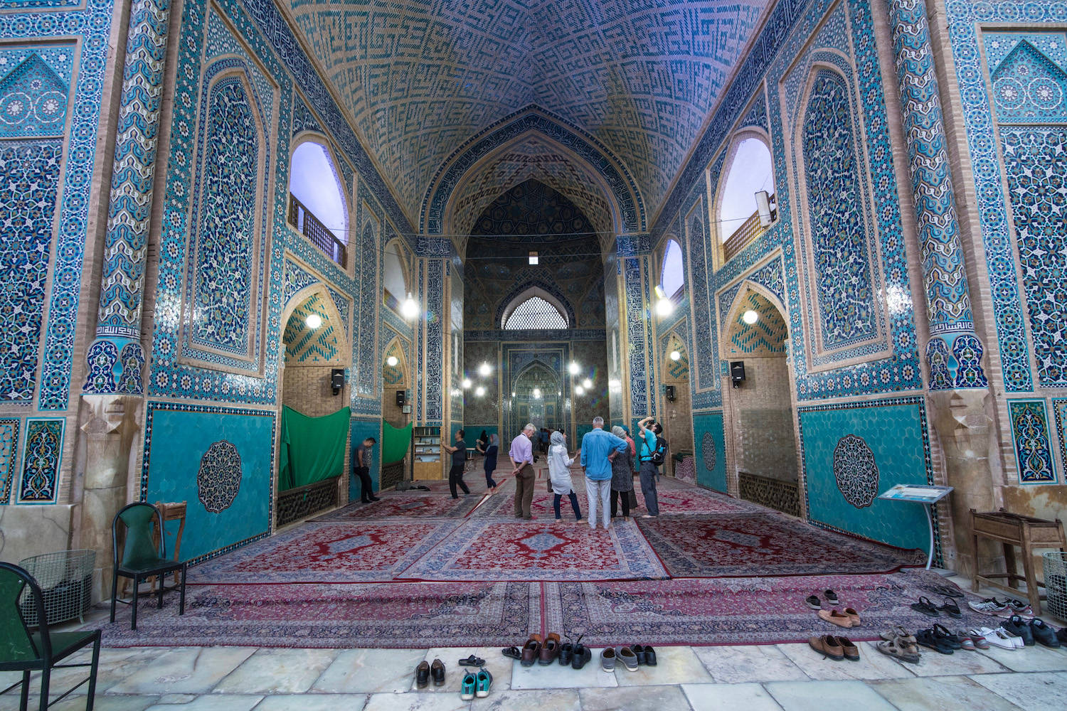 Iran 桌布 壁紙 — 宏偉的清真寺內部，有綠松石瓷磚牆壁、複雜的幾何圖案，以及遊客在華麗的紅色地毯上行走。