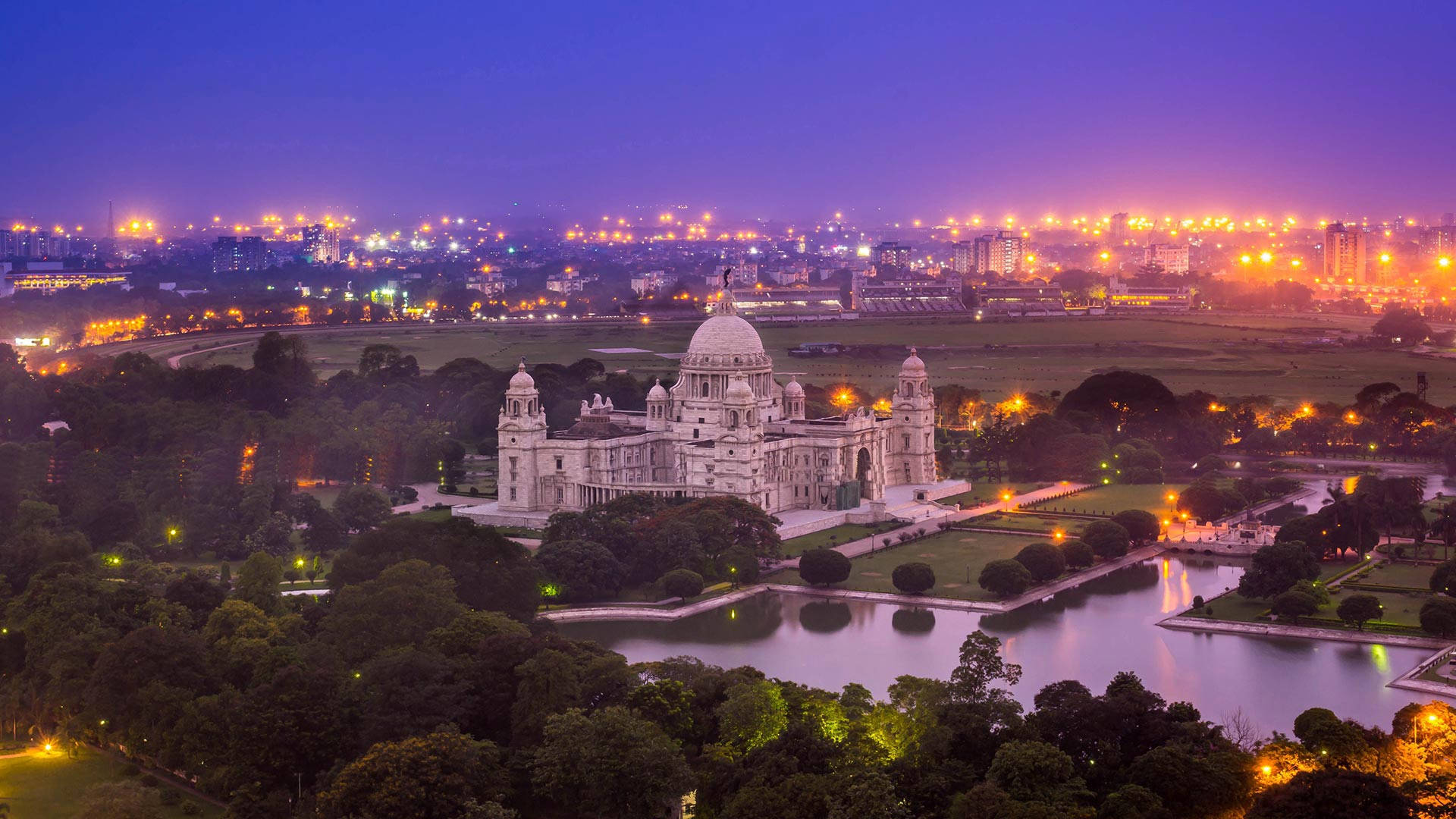 Kolkata 桌布 壁紙 — 宏偉的白色維多利亞紀念堂沐浴在暮色燈光中，周圍環繞著茂密的綠植和反射的水面，背景中城市天際線閃耀。
