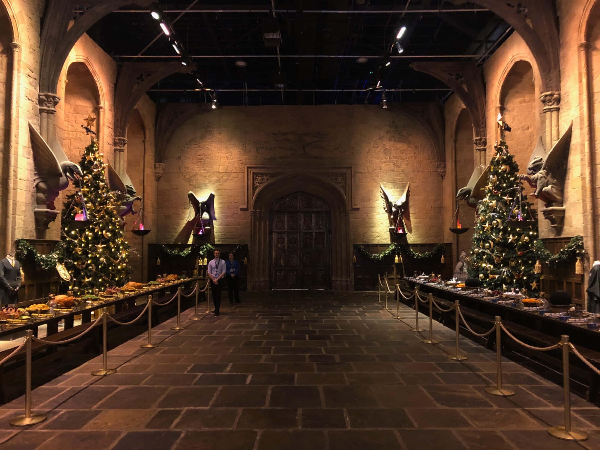 The Hogwarts Great Hall 桌布 壁紙 — 宏偉的石廳，高聳的拱門和節日聖誕樹在溫暖的聚光燈下排列在長長的宴會桌兩旁。