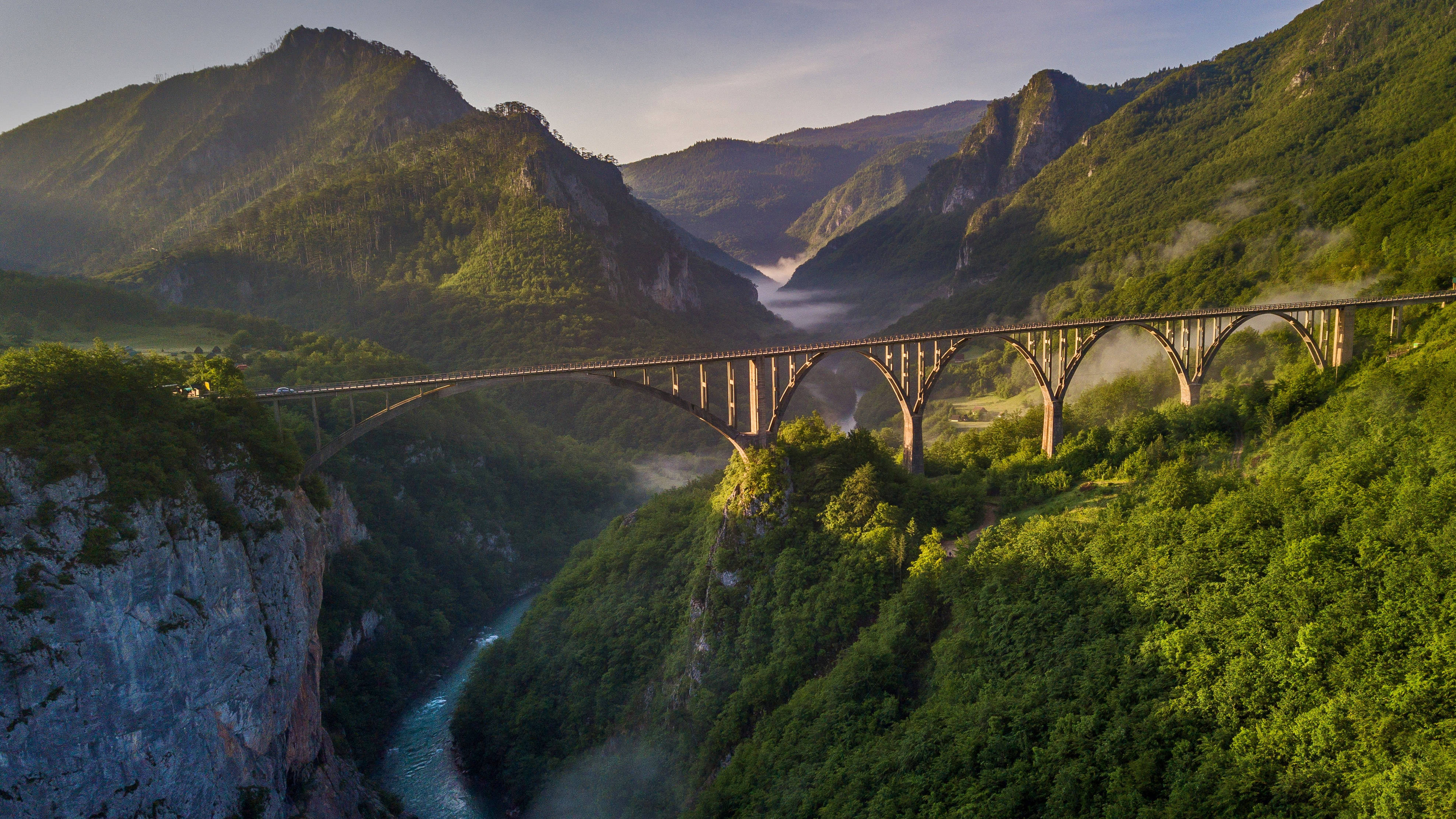 Montenegro 桌布 壁紙 — 宏偉的石拱橋橫跨深林峽谷，下方蜿蜒的河流在金色日出下閃耀。
