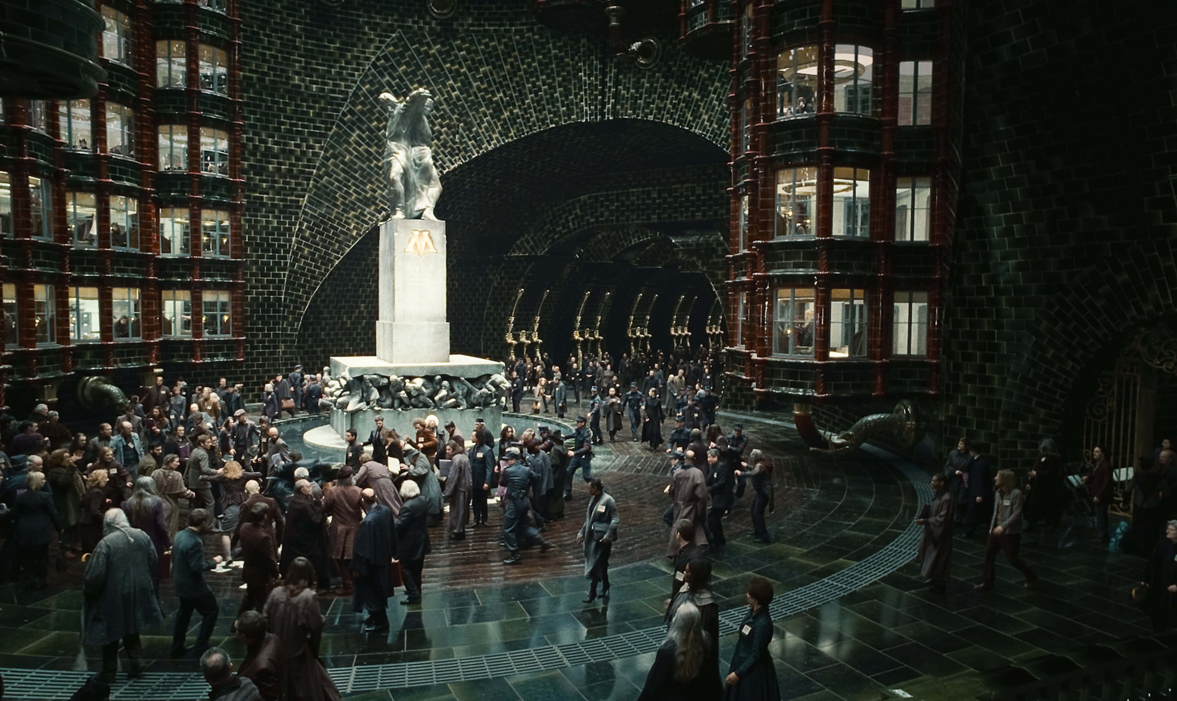 The Ministry Of Magic 桌布 壁紙 — 宏偉的石砌中庭，拱形天花板下透光的窗戶照亮人群圍繞中央雕像聚集。
