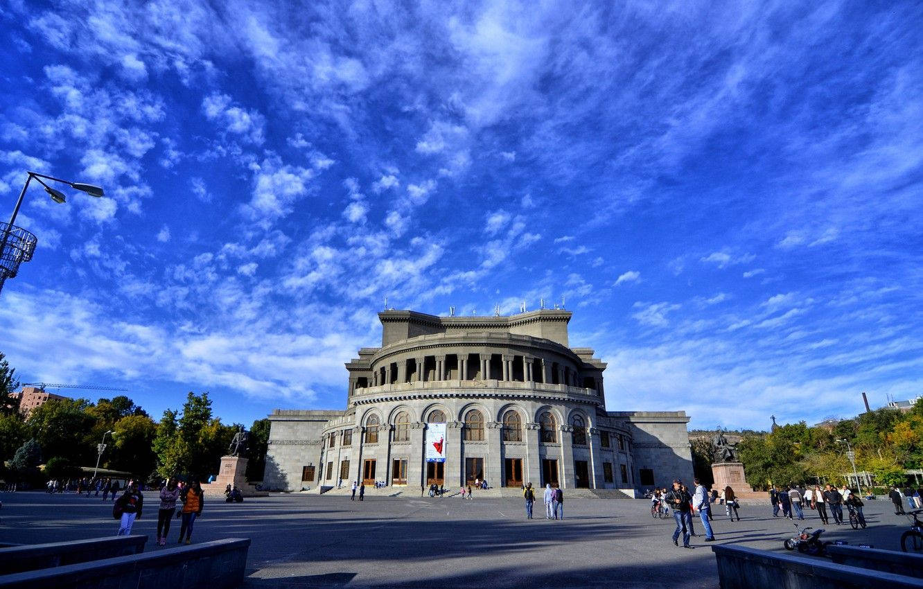 Armenia 桌布 壁紙 — 宏偉的石造劇院帶有拱形窗戶，矗立在充滿活力的藍天下，有縷縷白雲，人們在廣場上漫步。