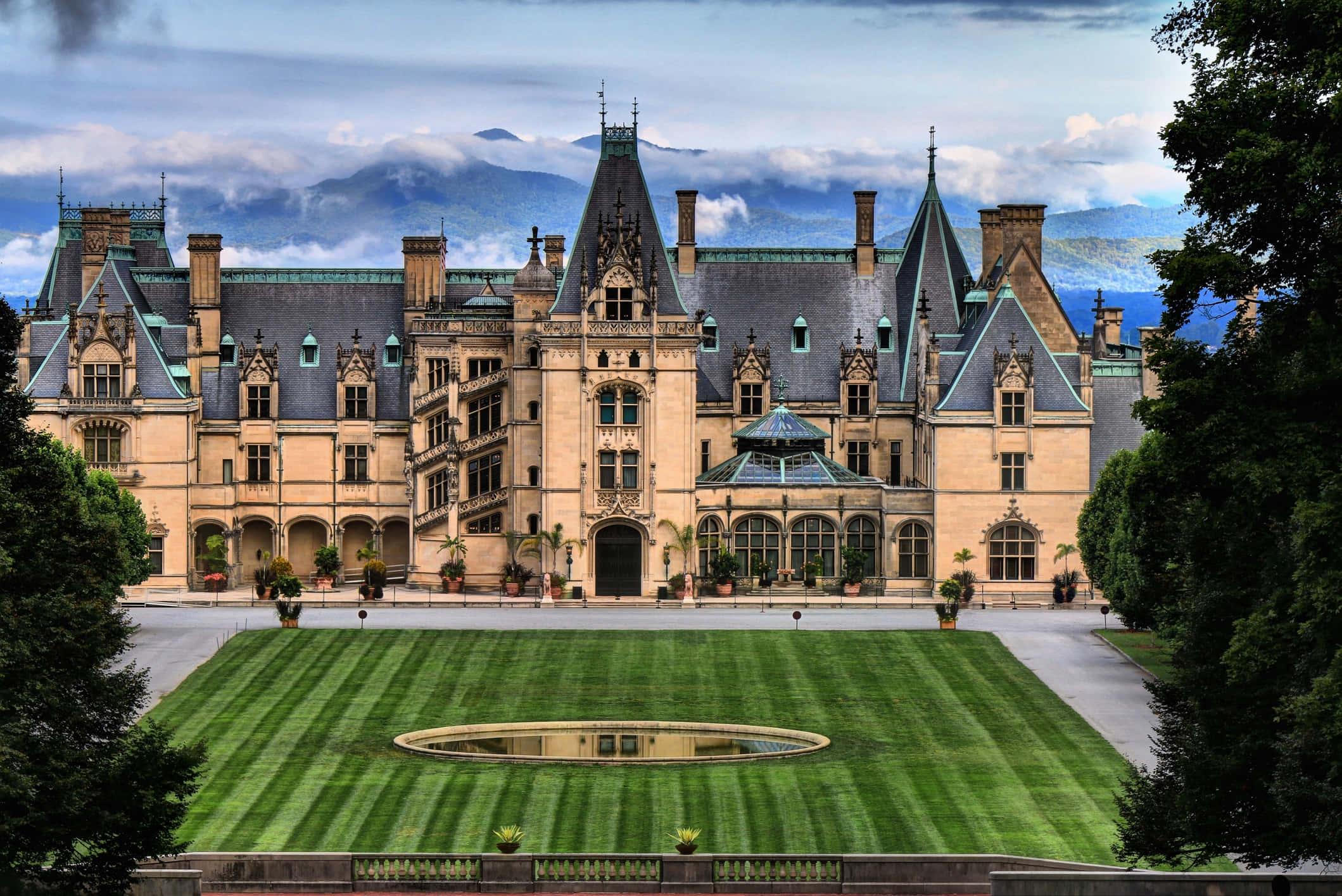Biltmore Estate 桌布 壁紙 — 宏偉的石頭城堡，擁有陡峭的山牆屋頂和角樓，矗立在修剪整齊的草坪前，中央有倒影池，天空多雲。