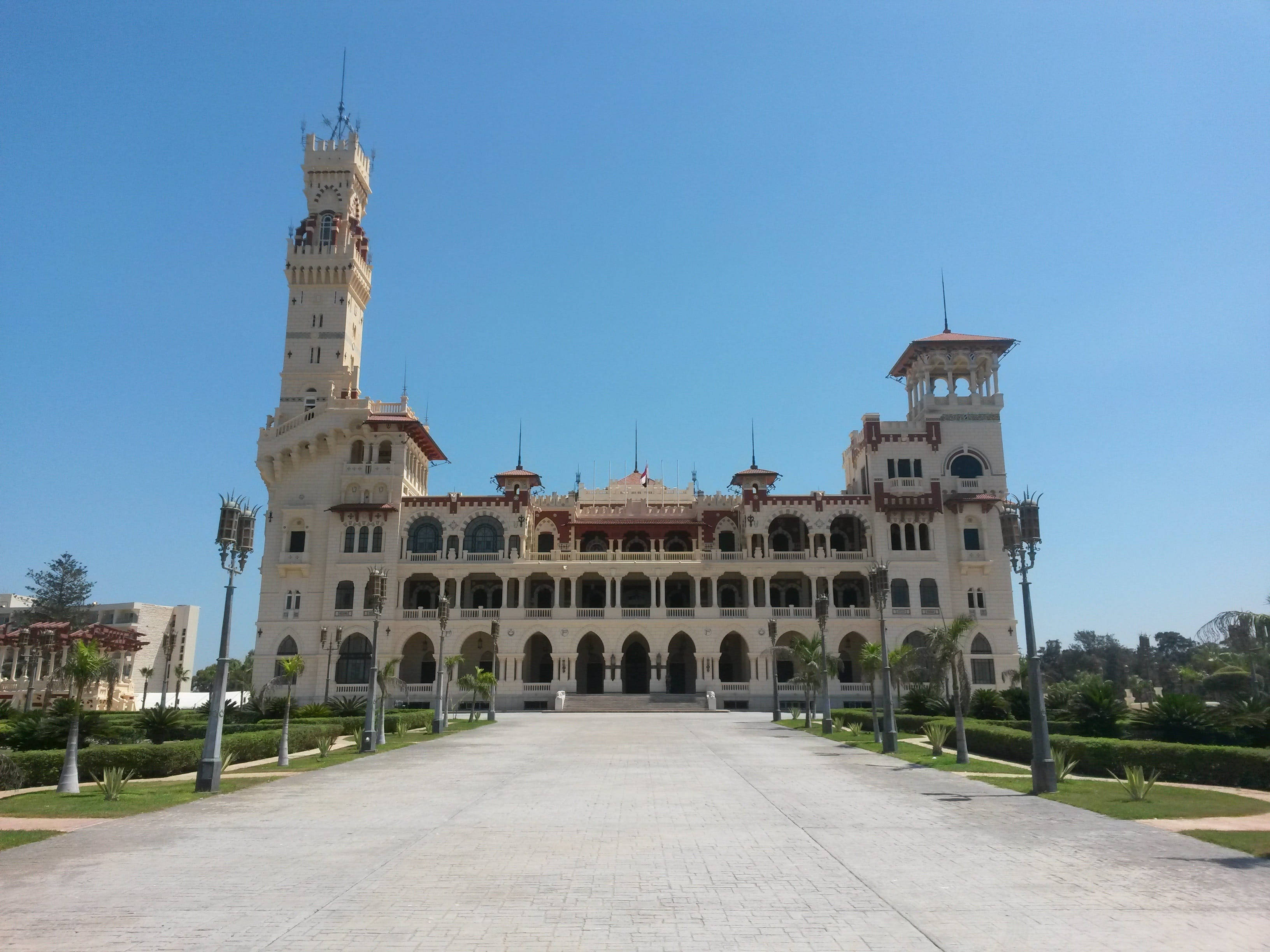 Alexandria 桌布 壁紙 — 宏偉的米色宮殿，擁有華麗的塔樓和拱形陽台，矗立在晴朗的藍天下，兩旁種滿棕櫚樹和修剪整齊的花園。