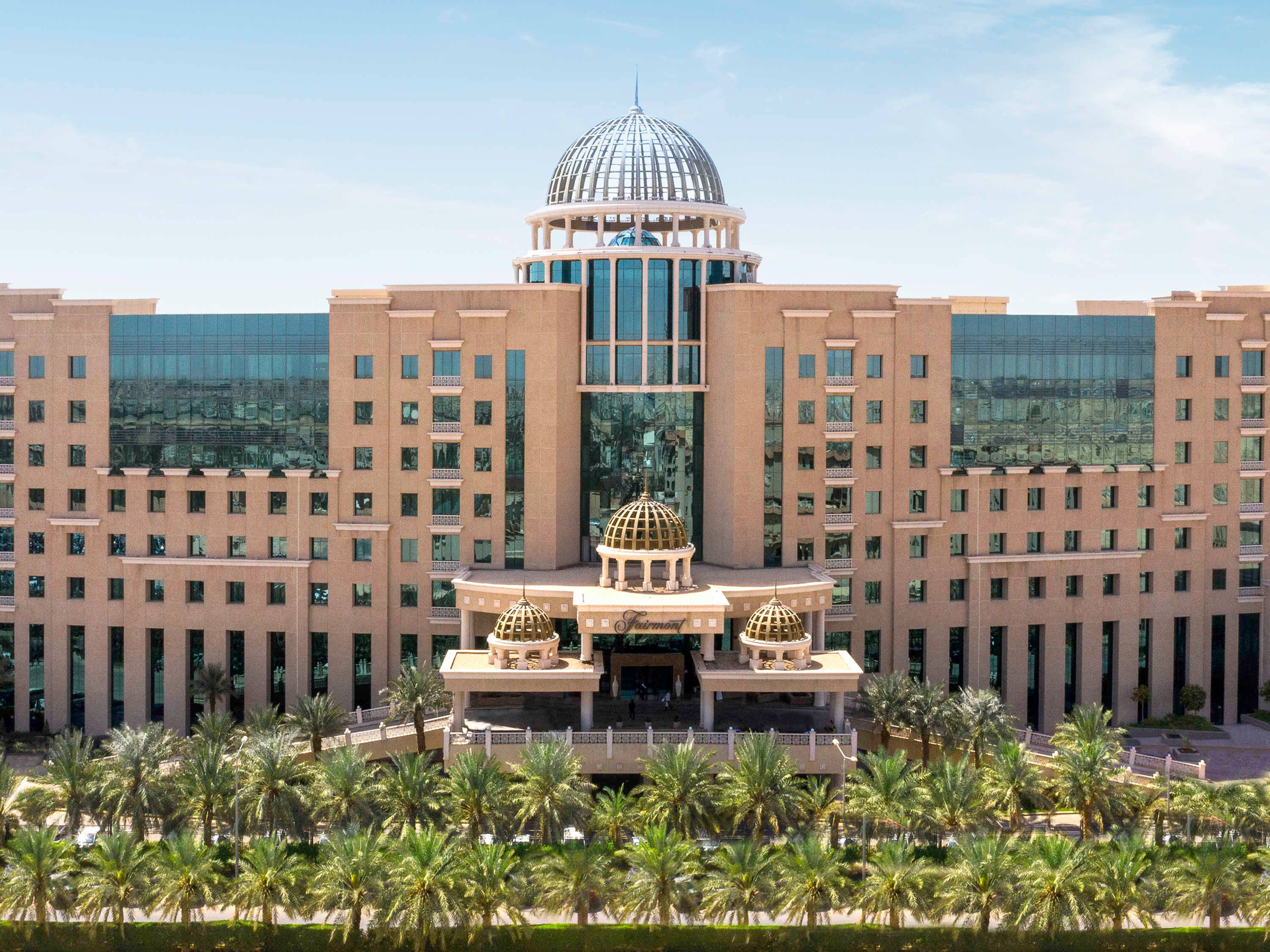 Riyadh 桌布 壁紙 — 宏偉的米色酒店，擁有玻璃圓頂和金色裝飾，周圍環繞著茂密的棕櫚樹，在明亮的藍天下。