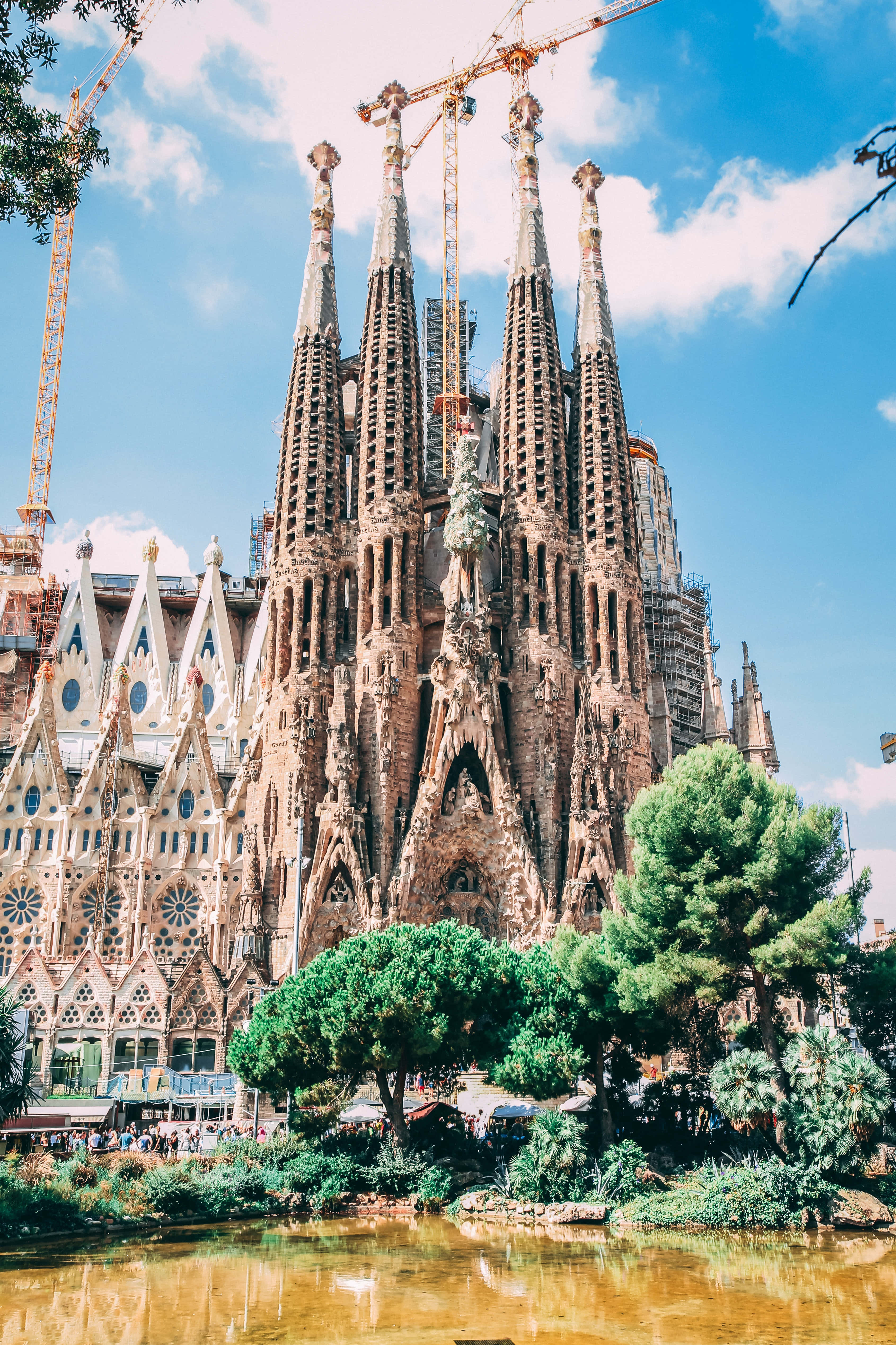 Barcelona 桌布 壁紙 — 宏偉的聖家堂大教堂，尖塔在藍天下，周圍環繞著茂密的綠樹和倒映的池塘。
