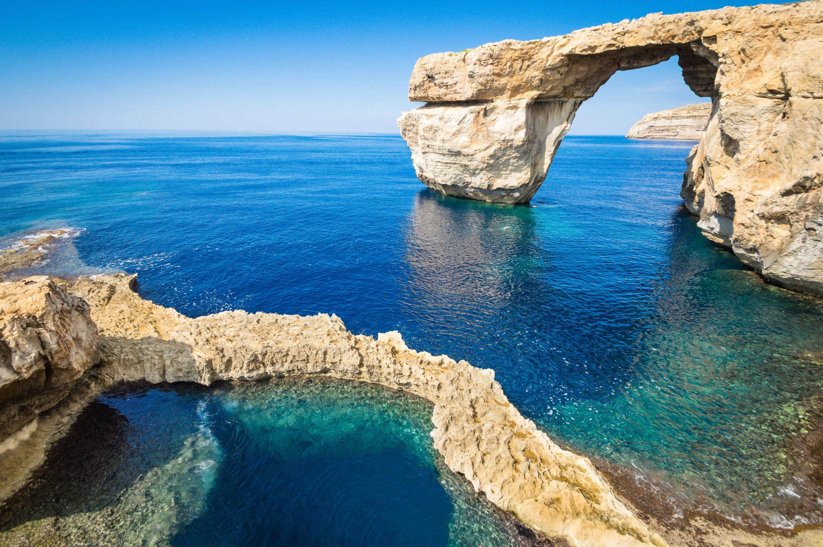 Malta 桌布 壁紙 — 宏偉的自然岩石拱門從碧綠的海水中升起，在清澈的藍天下，下方是平靜的大海。