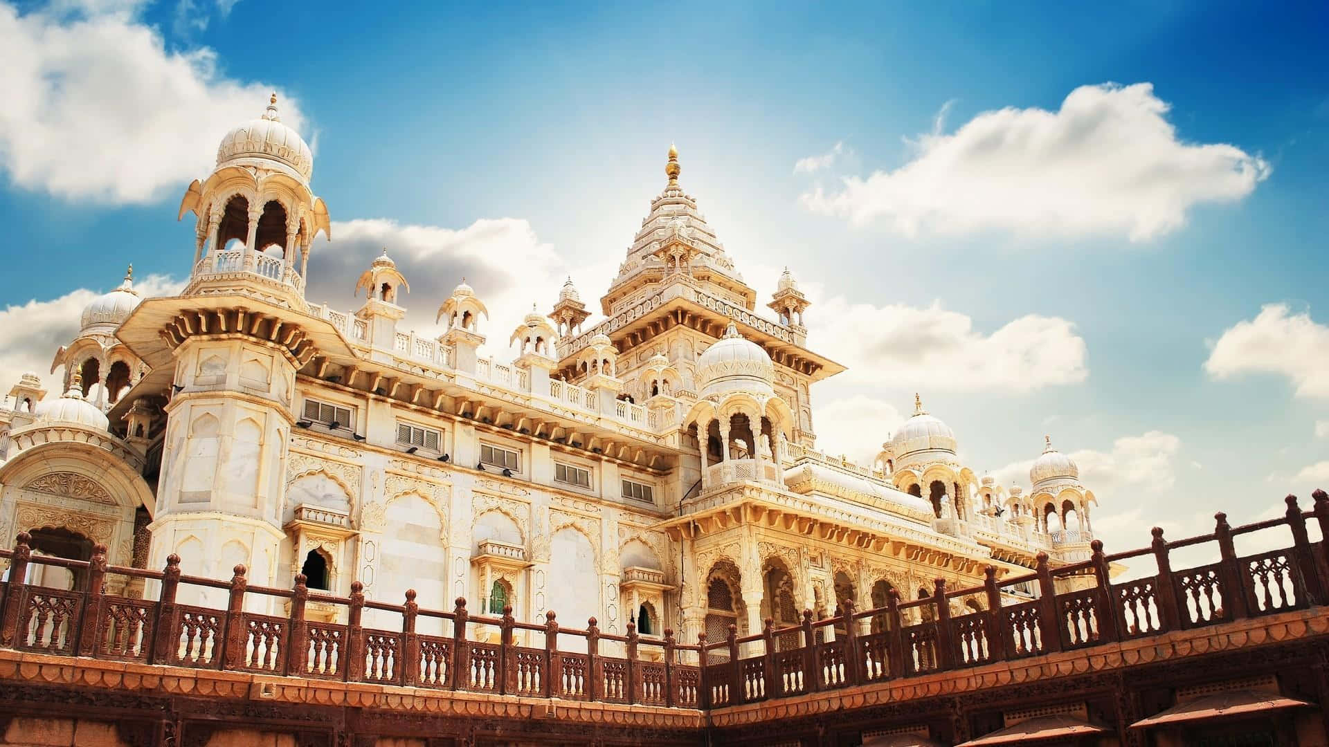 Rajasthan 桌布 壁紙 — 宏偉的象牙色宮殿，擁有圓頂和宣禮塔，沐浴在金色陽光下，背景是晴朗的藍天和蓬鬆的白雲。