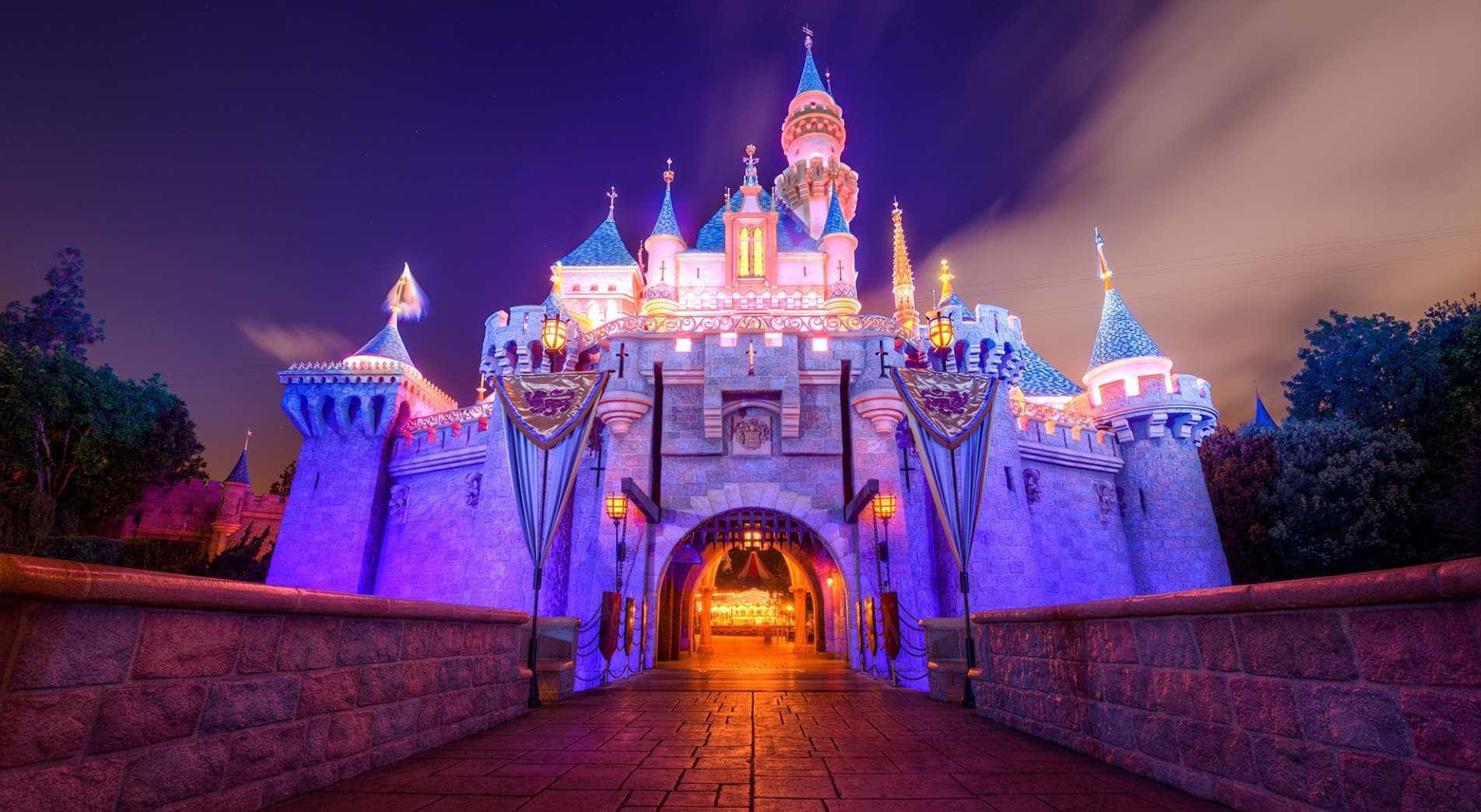 Disney Castle 桌布 壁紙 — 宏偉的迪士尼城堡在夜晚被紫色和金色燈光照亮，一條石橋通向發光的入口。