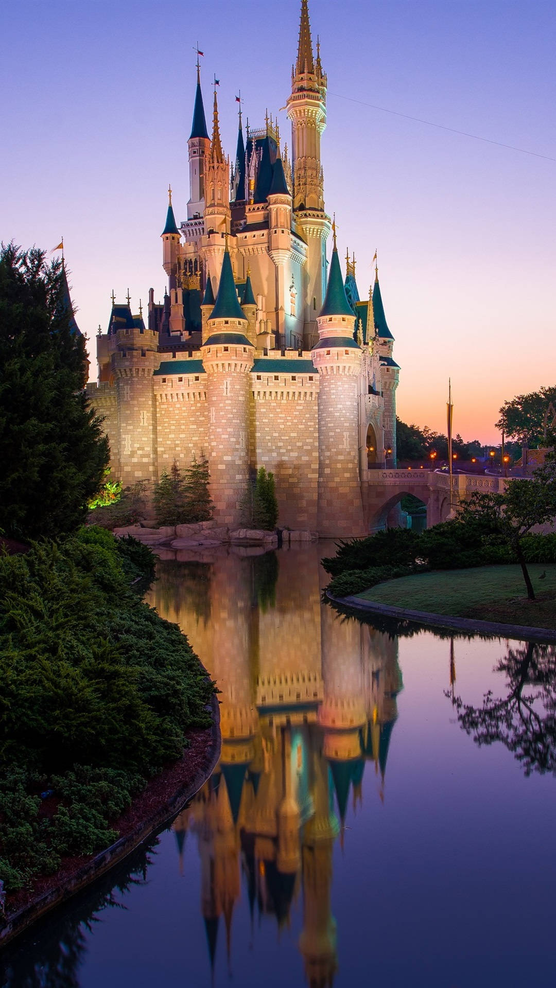Disney Castle 桌布 壁紙 — 宏偉的迪士尼城堡在黃昏時分被照亮，其尖塔和城樓完美地倒映在下方平靜的水面上。