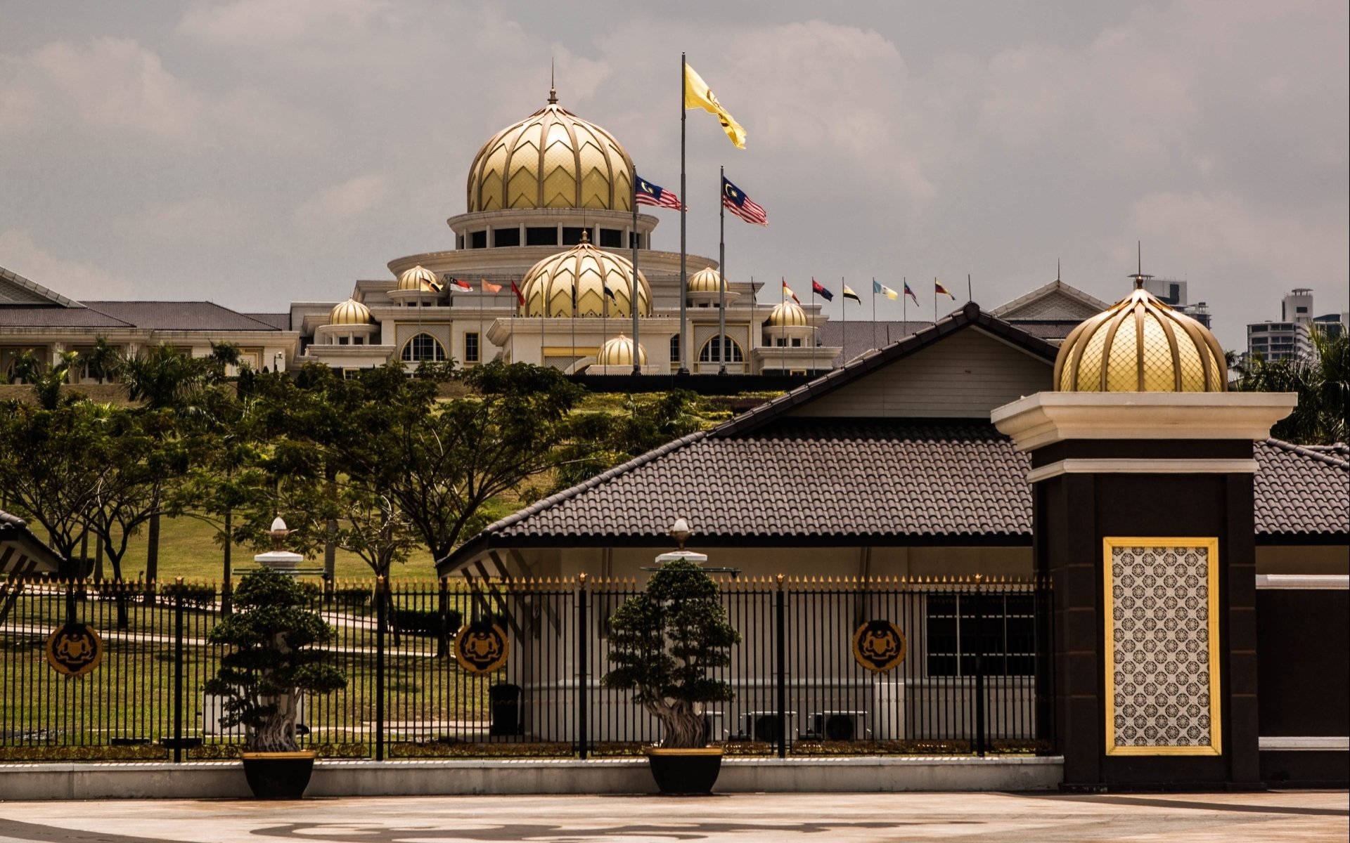 Jakarta 桌布 壁紙 — 宏偉的金色圓頂清真寺，旗幟飄揚，矗立在黑色鐵門和修剪整齊的綠植之後，天空多雲。