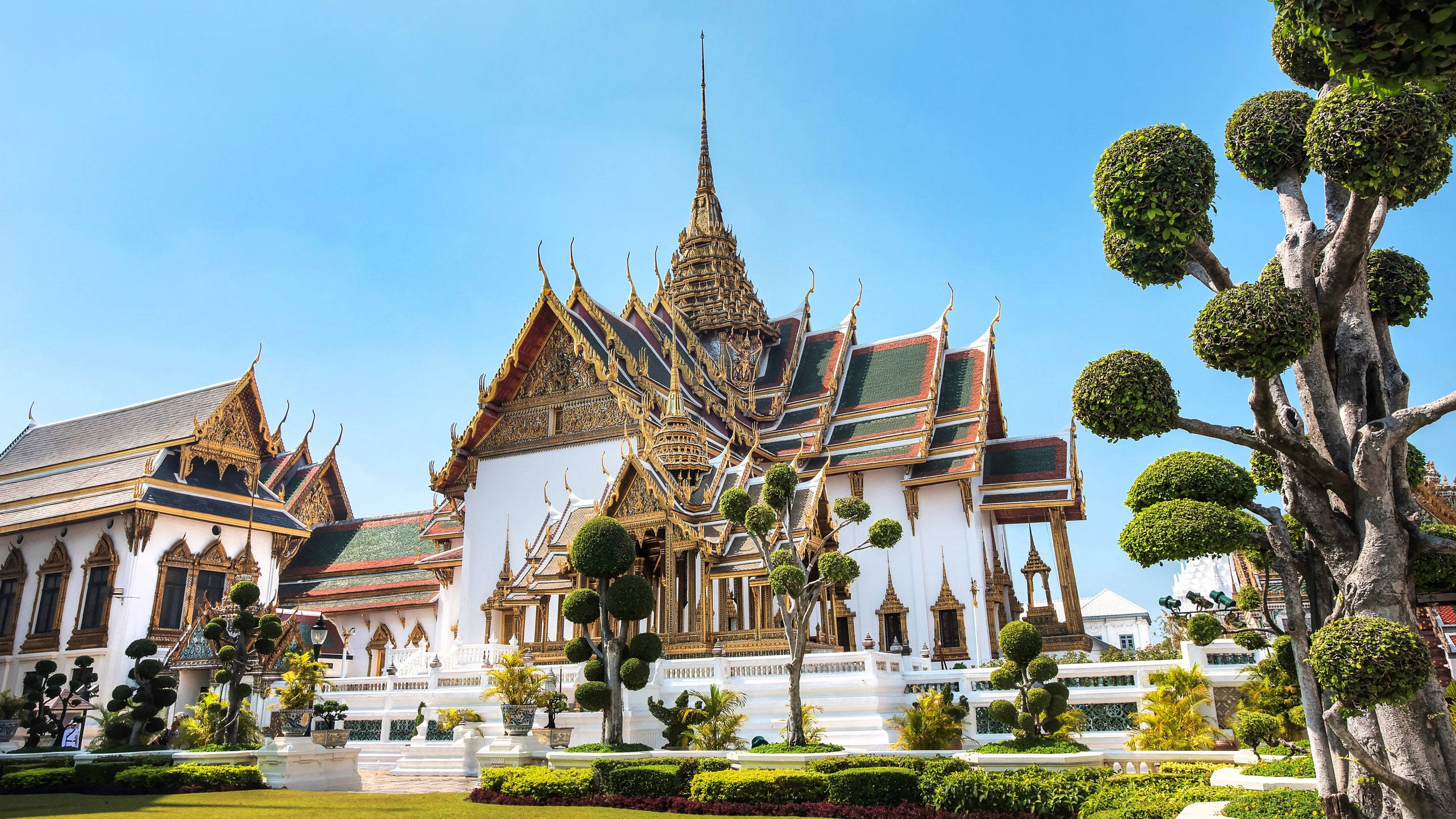 Bangkok 桌布 壁紙 — 宏偉的金色寺廟，擁有精緻的尖頂和修剪整齊的綠樹，在清澈的藍天下。