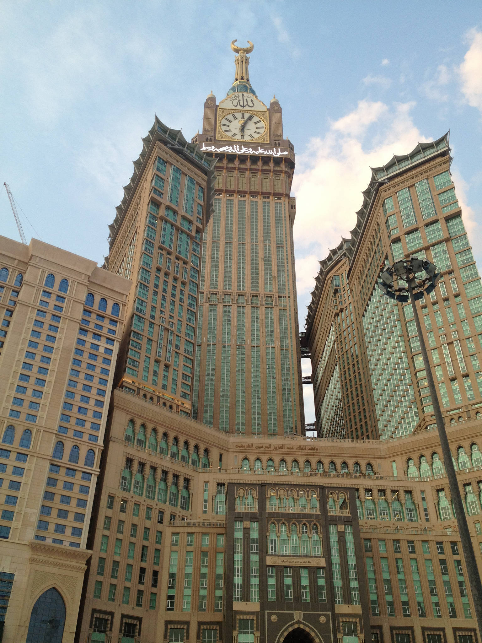 Saudi 桌布 壁紙 — 宏偉的鐘樓，頂部有金色新月，矗立於高聳的米色建築之上，周圍是現代玻璃摩天大樓，天空部分多雲。