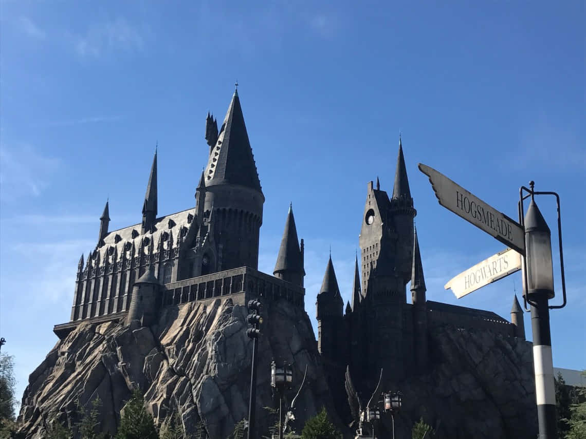 Hogsmeade 桌布 壁紙 — 宏偉的霍格華茲城堡坐落在岩石懸崖上，藍天白雲下，路牌指向活米村和霍格華茲。