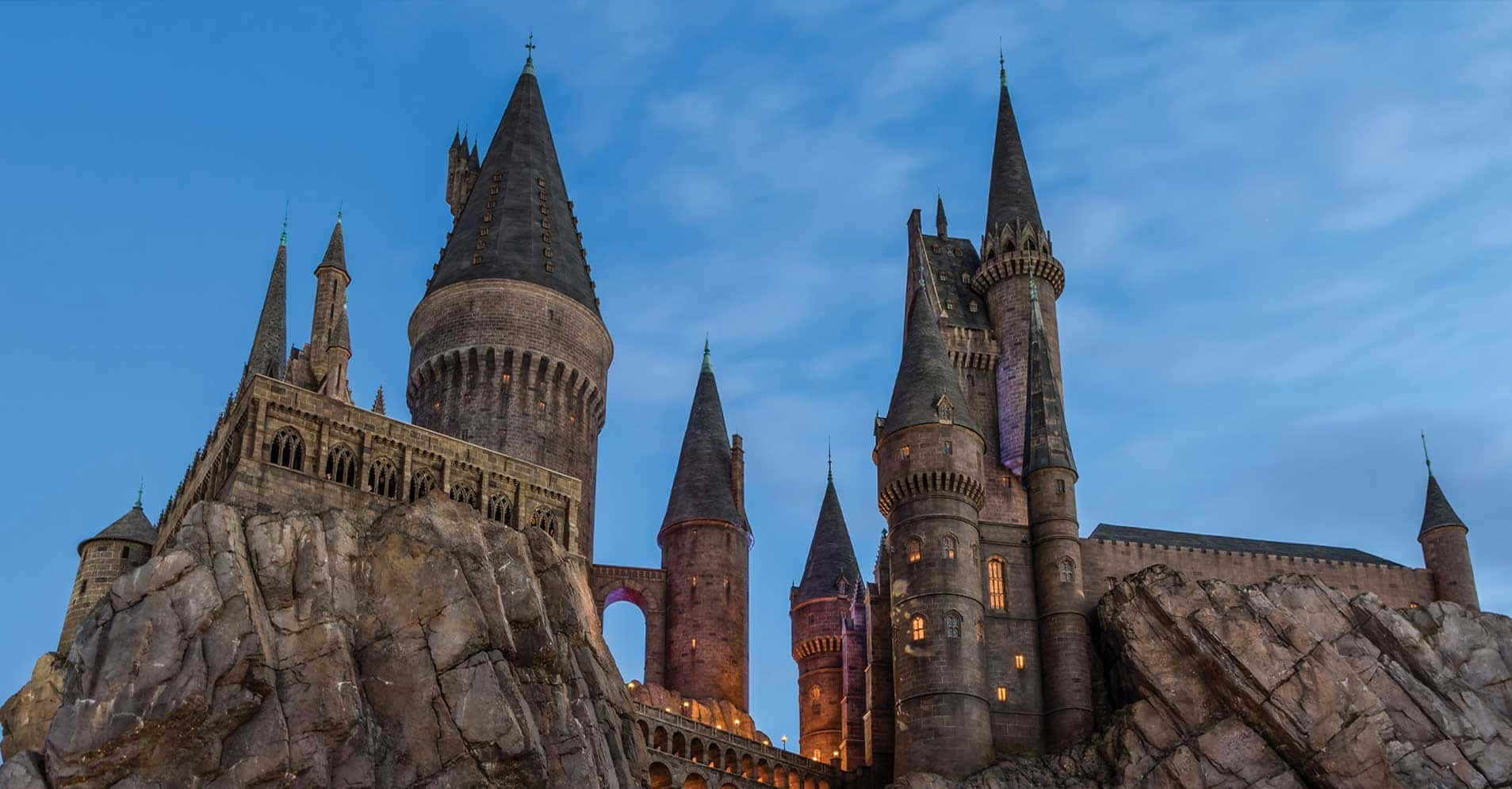 Hogsmeade 桌布 壁紙 — 宏偉的霍格華茲城堡矗立在岩石懸崖上，在暮色天空下閃爍著窗戶的光芒。