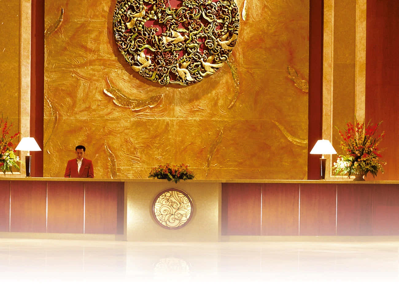 Hotel 桌布 壁紙 — 宏偉的飯店大廳，配有金色華麗牆飾、身穿紅色西裝的員工，以及在溫暖燈光下的花卉佈置。