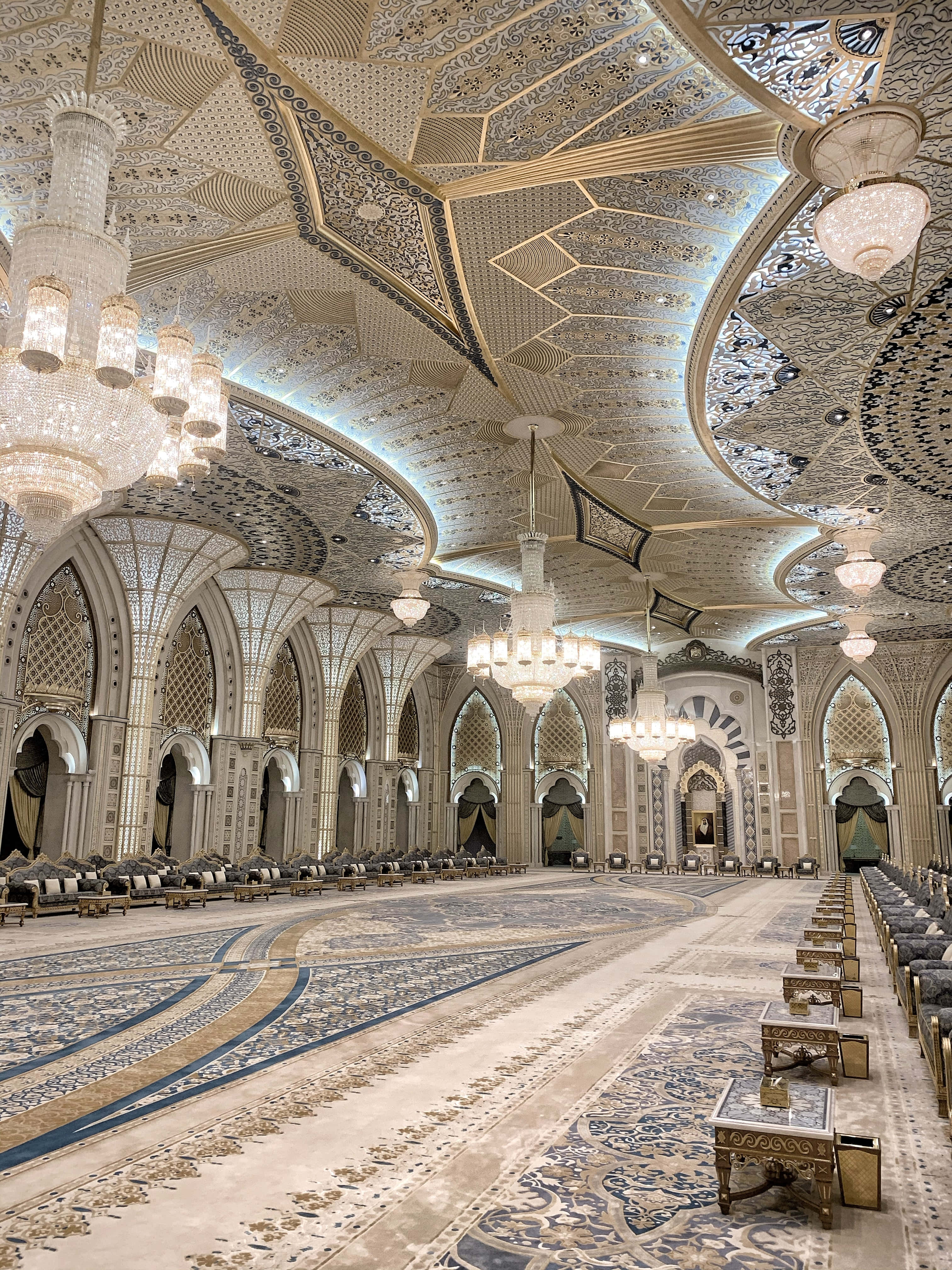 Ballroom 桌布 壁紙 — 宏偉華麗的大廳，帶有複雜的幾何天花板圖案、金色點綴和吊燈，照亮一排排華麗的座位。