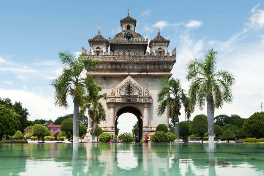Laos 桌布 壁紙 — 宏偉華麗的拱門，帶有精緻雕刻，雄偉地矗立在平靜的倒影池後方，兩旁種植棕櫚樹，上方是明亮的藍天。