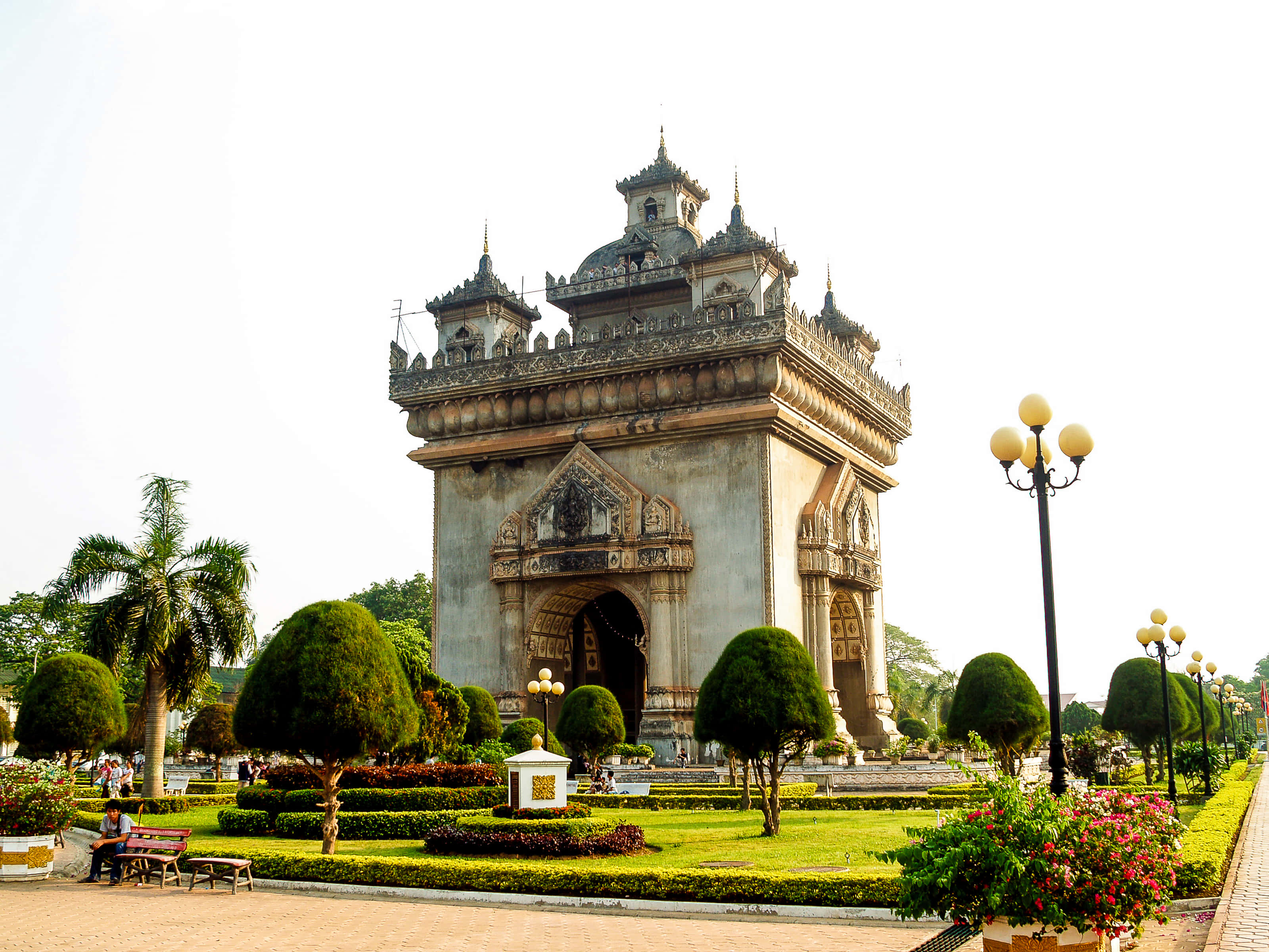 Laos 桌布 壁紙 — 宏偉華麗的拱門，雕刻精緻，矗立在修剪整齊的花園中，棕櫚樹和盛開的花朵在明亮的日光下。