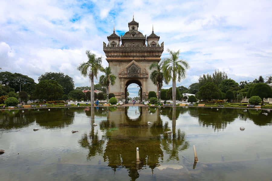 Laos 桌布 壁紙 — 宏偉華麗的拱門倒映在平靜的水面上，周圍環繞著棕櫚樹和茂密的綠植，天空部分多雲。
