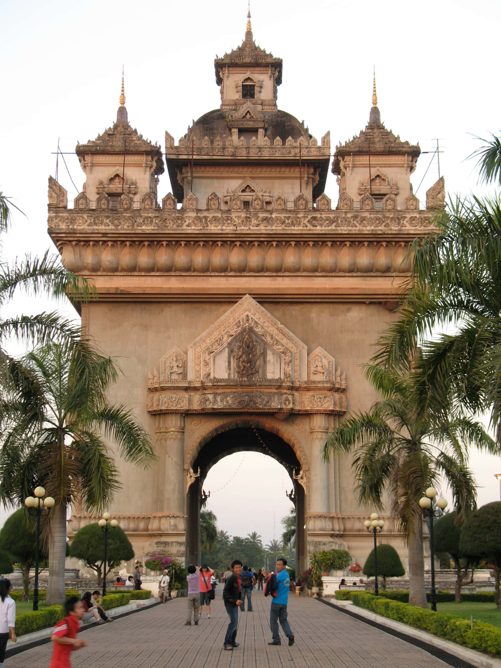 Laos 桌布 壁紙 — 宏偉華麗的石拱門，帶有精緻雕刻和金色尖頂，以棕櫚樹和鋪砌步道上的遊客為背景。