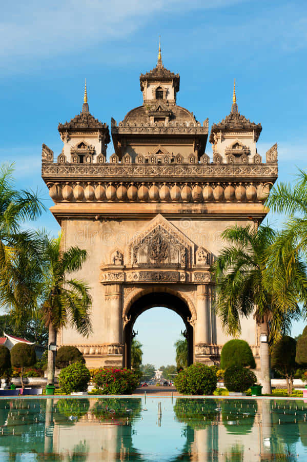 Laos 桌布 壁紙 — 宏偉華麗的石拱門，帶有精緻雕刻和金色尖頂，倒映在清澈藍天下寧靜的水池中。
