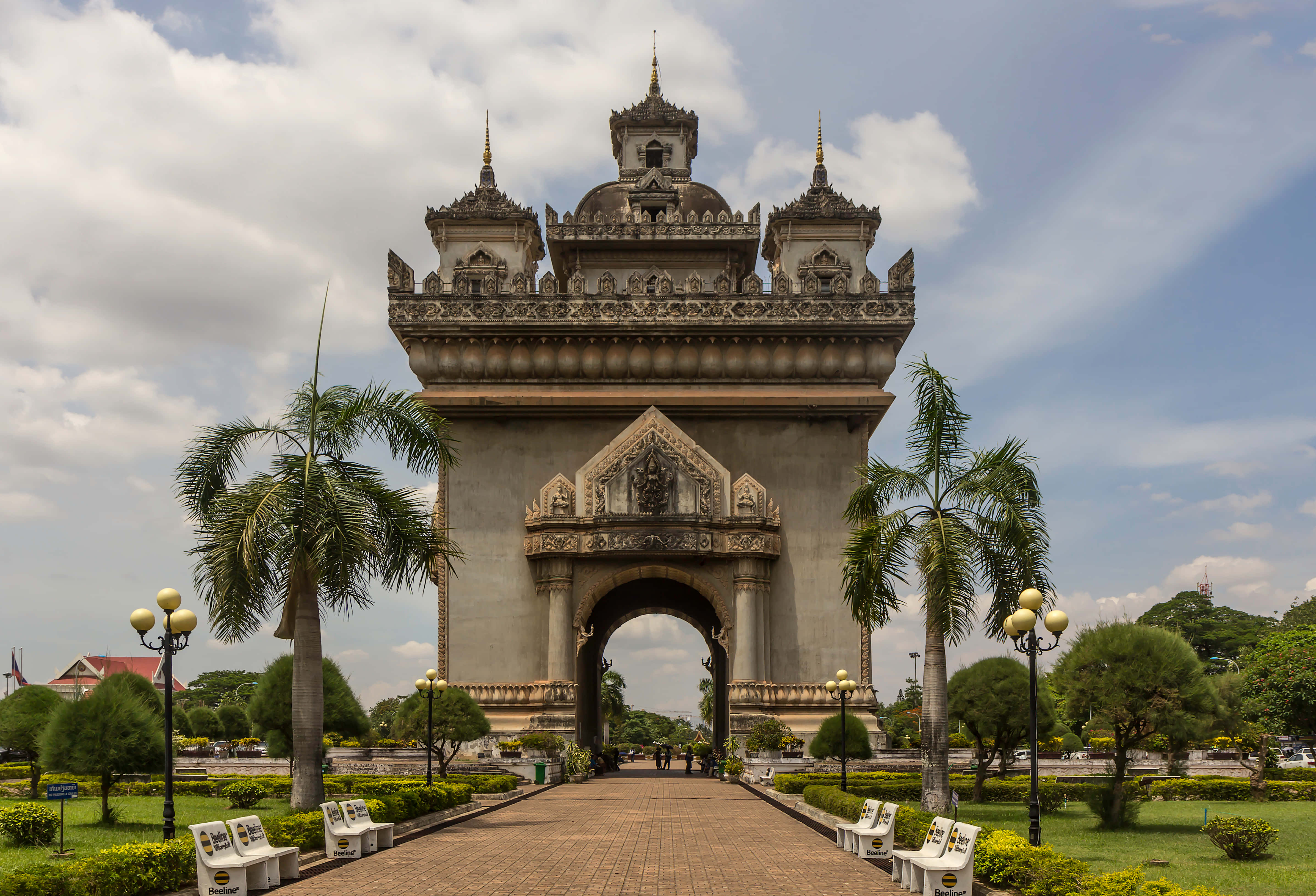 Laos 桌布 壁紙 — 宏偉華麗的石拱門，雕刻精細，兩旁種有棕櫚樹，天空部分多雲。