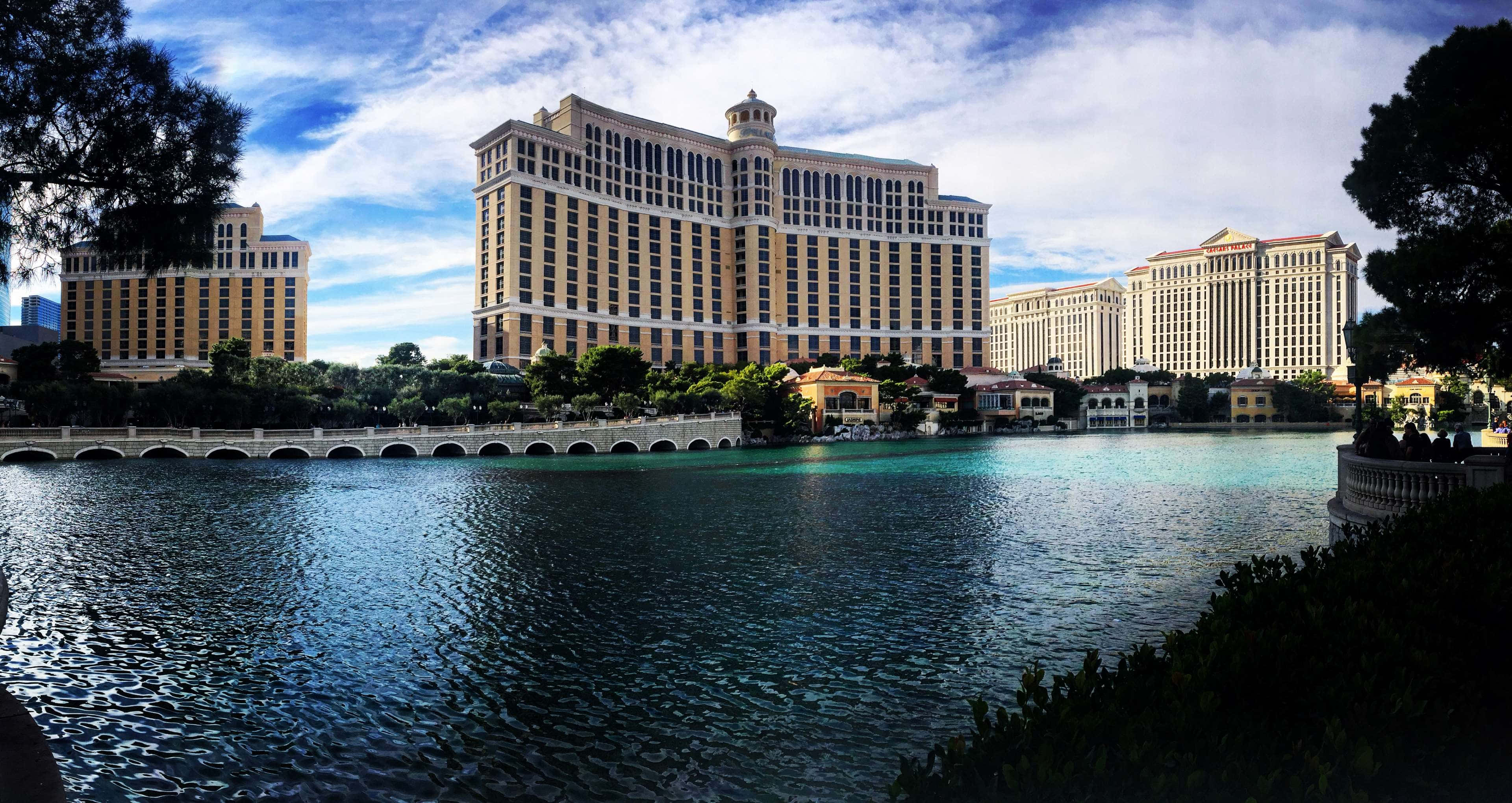 4K Las Vegas 桌布 壁紙 — 宏偉豪華酒店塔樓在晴朗藍天與零星白雲下，矗立於平靜的藍綠色運河後方。