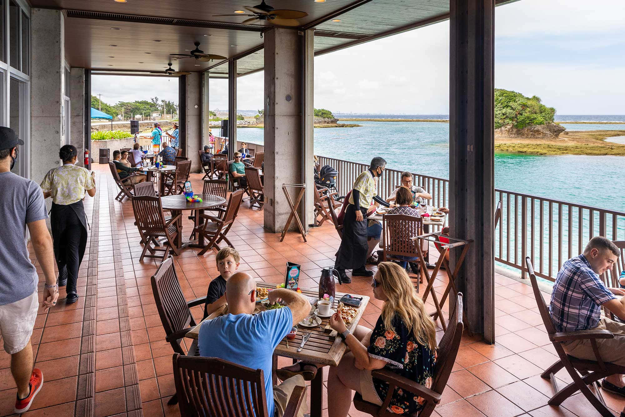Seaside 桌布 壁紙 — 客人在有蓋天井下的戶外餐桌用餐，俯瞰著碧綠的海水和遠處的綠色島嶼。