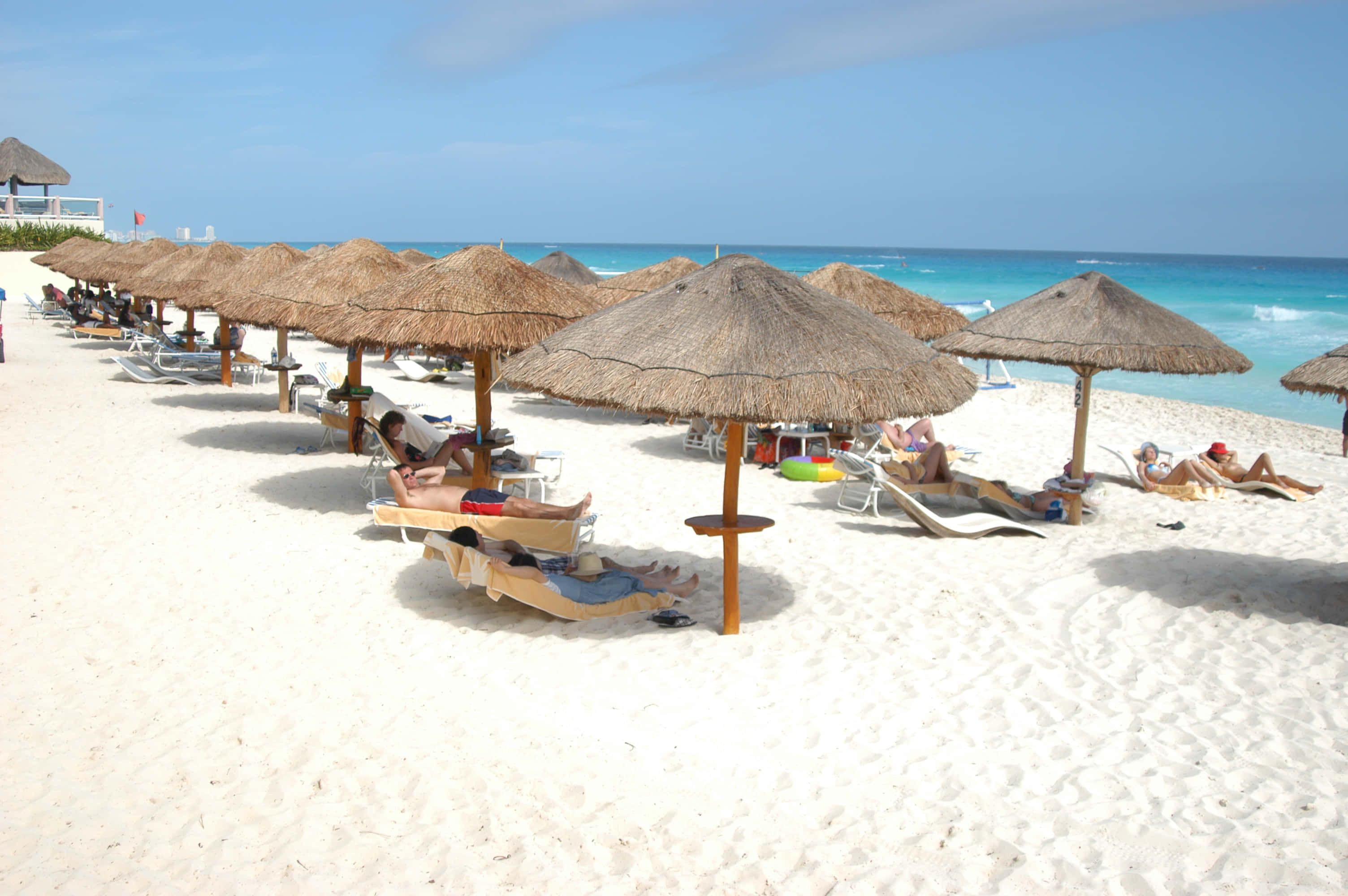 Mexican Beach 桌布 壁紙 — 客人在清澈藍天下，在原始白沙灘上，在稻草傘下放鬆，旁邊是碧綠的海浪。