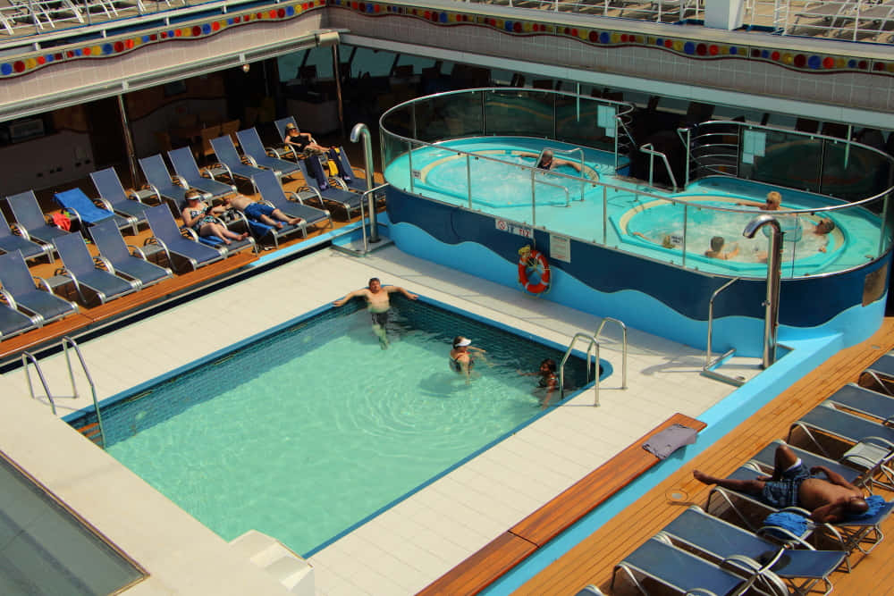 Carnival Conquest 桌布 壁紙 — 客人在陽光明媚的郵輪甲板上，在周圍擺放著躺椅的明亮藍色泳池中放鬆和游泳。