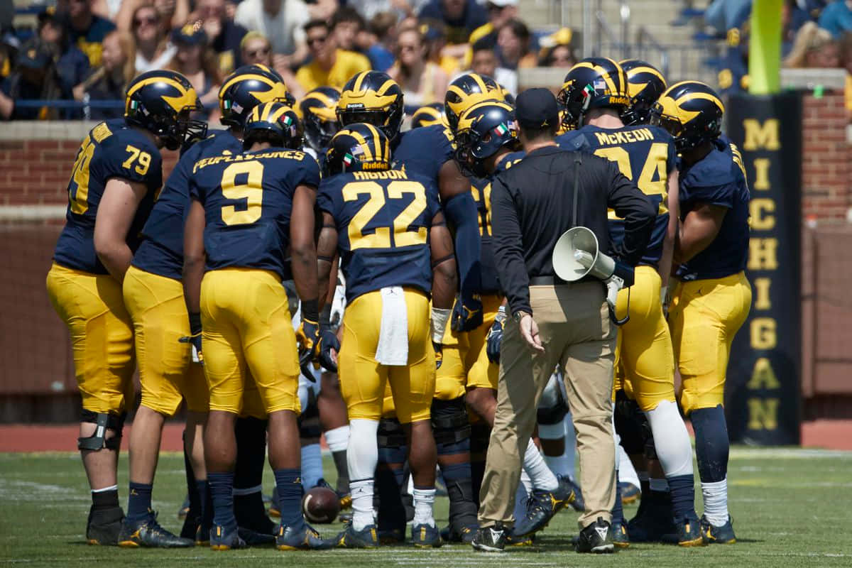 Michigan Football 桌布 壁紙 — 密西根大學美式足球員身穿藍黃色球衣，在陽光明媚的球場上圍成一圈，教練手持擴音器。