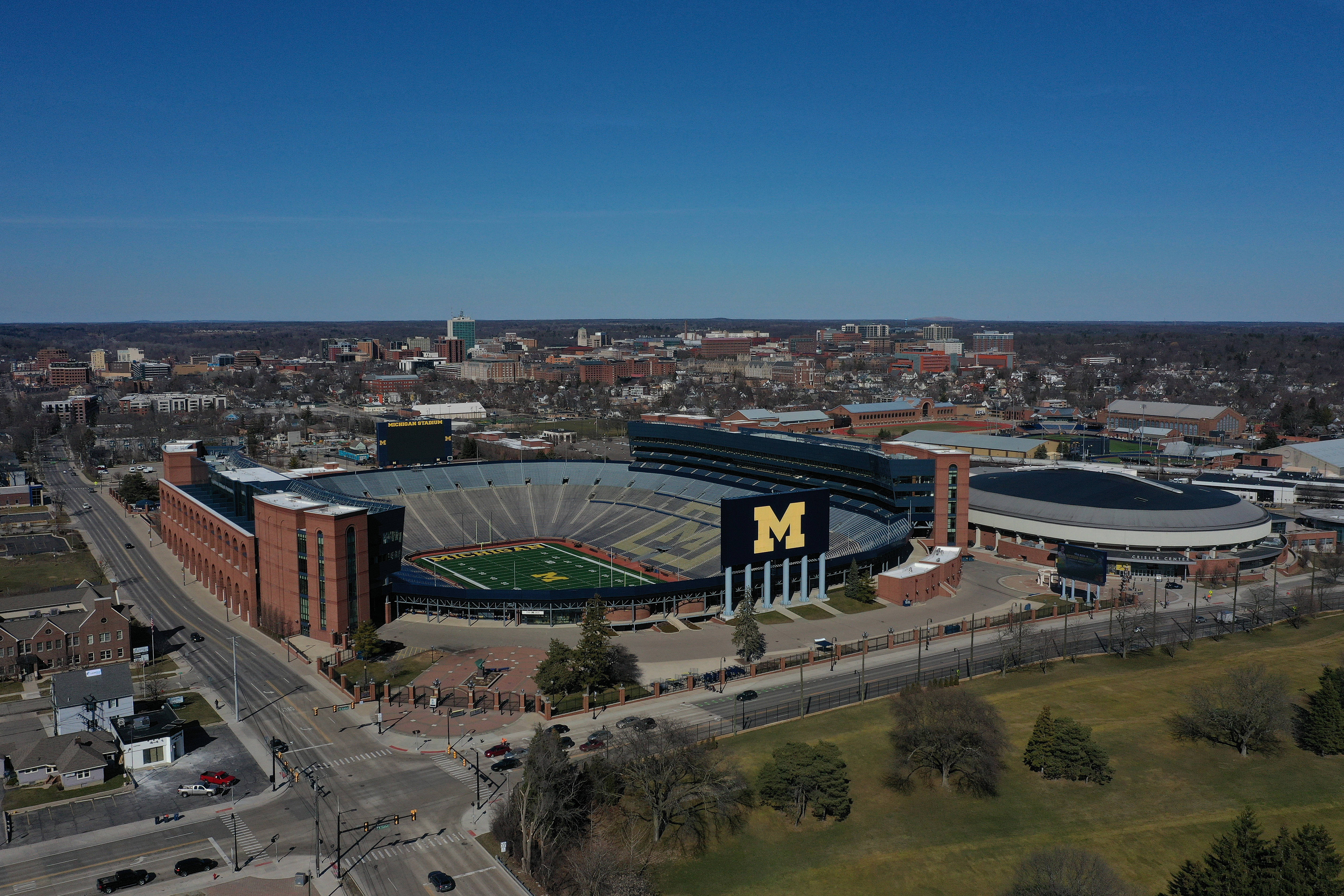 學校 桌布 壁紙 — 密西根體育場的鳥瞰圖，其標誌性的「M」標誌在晴朗的藍天下，被城市建築和綠色田野包圍。