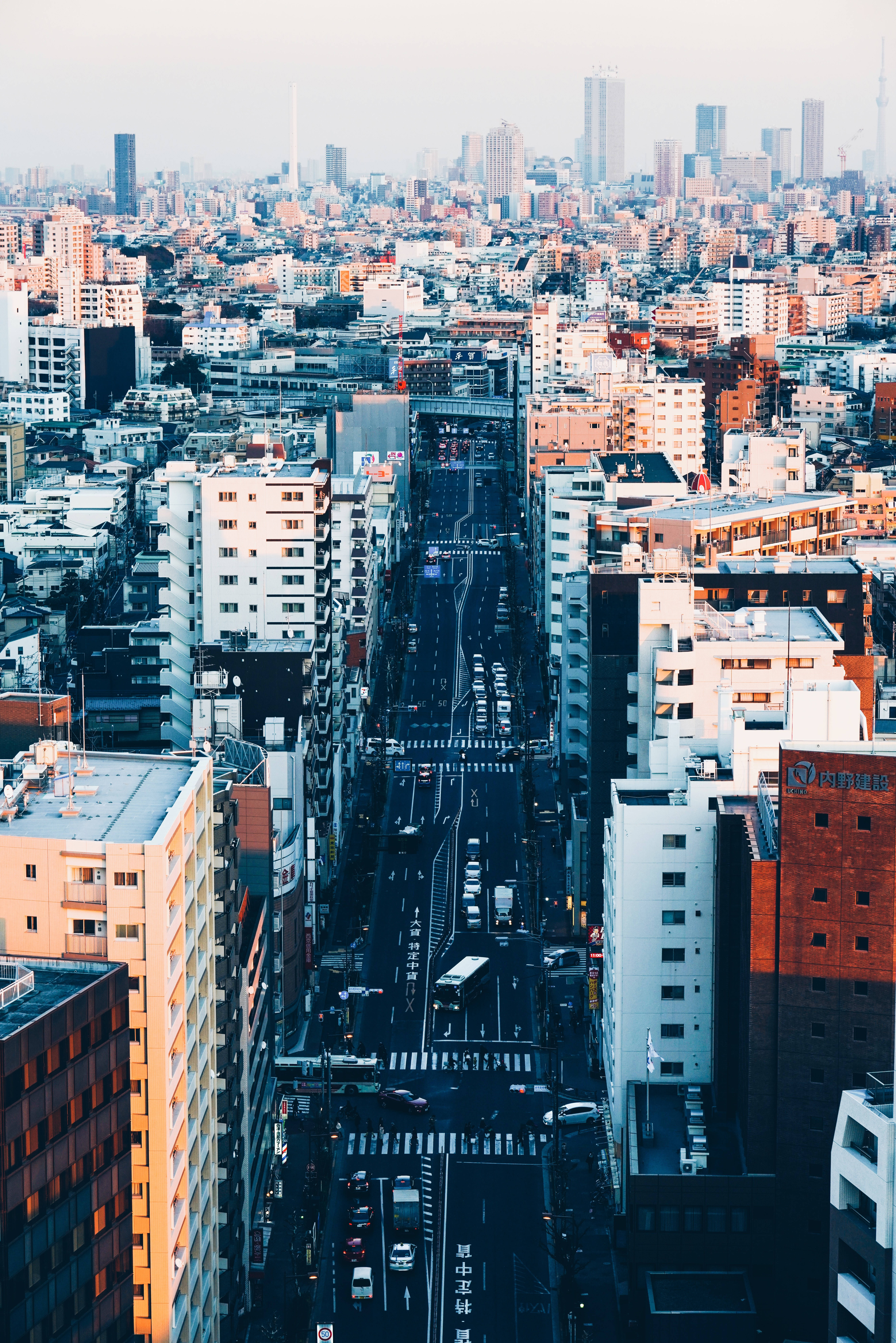 東京 桌布 壁紙 — 密集城市峽谷的鳥瞰圖，高聳的建築物和一條繁忙的街道延伸至朦朧的城市景觀。