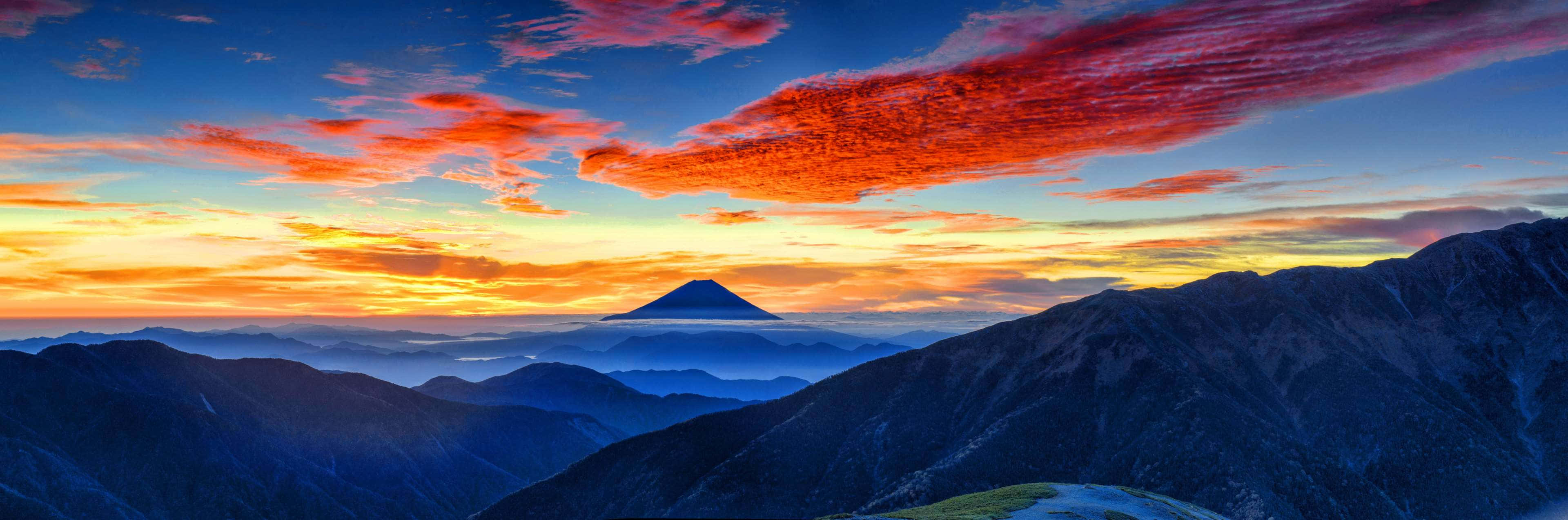 Mountain Landscape 桌布 壁紙 — 富士山在橙紅色雲彩點綴的熾熱日落天空下，於層疊的藍色山脈之上寧靜地聳立。