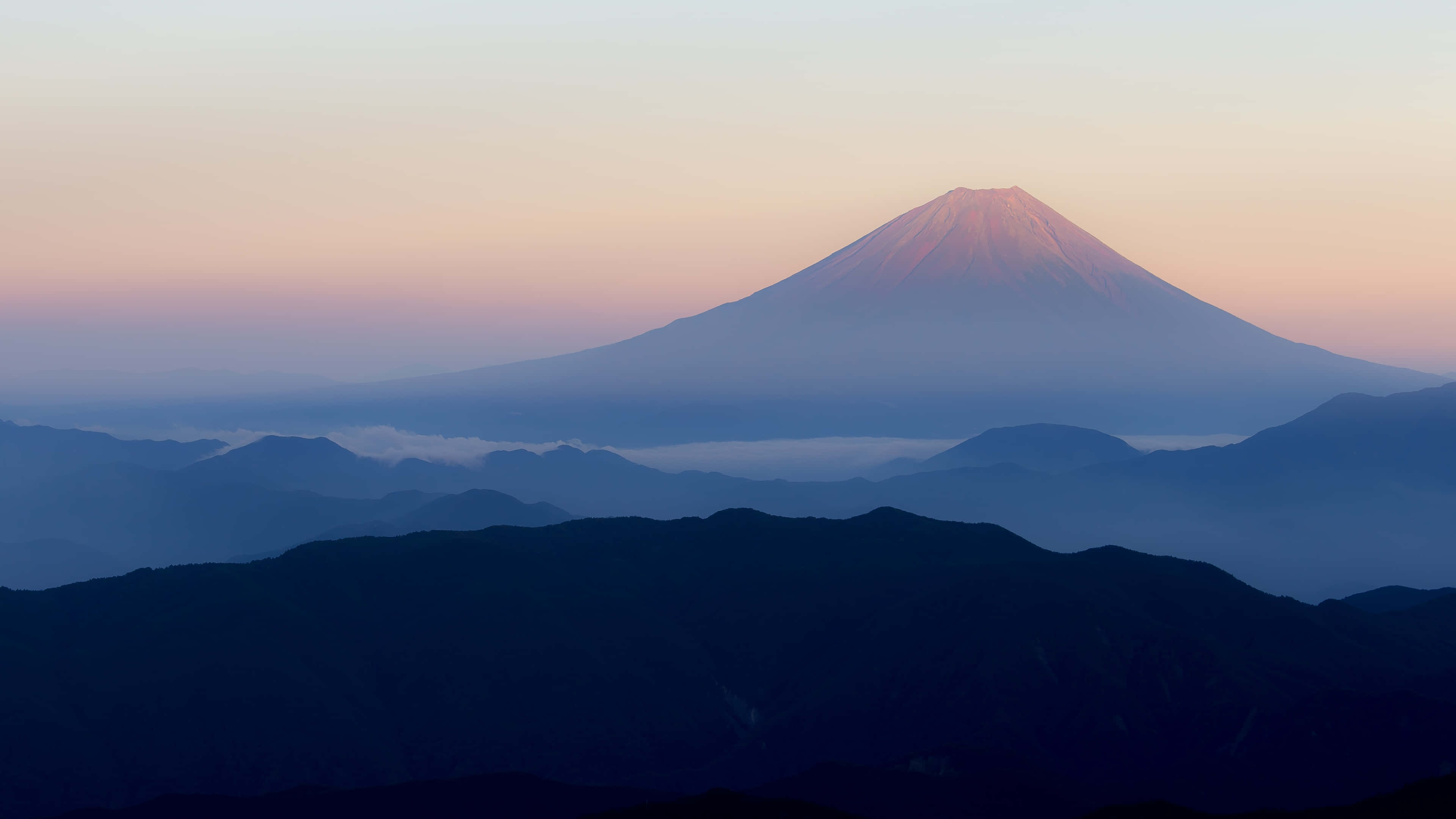 日本 桌布 壁紙 — 富士山在黎明時分雄偉地升起，柔和的粉紅色和藍色調在層疊的山巒輪廓之上描繪天空。