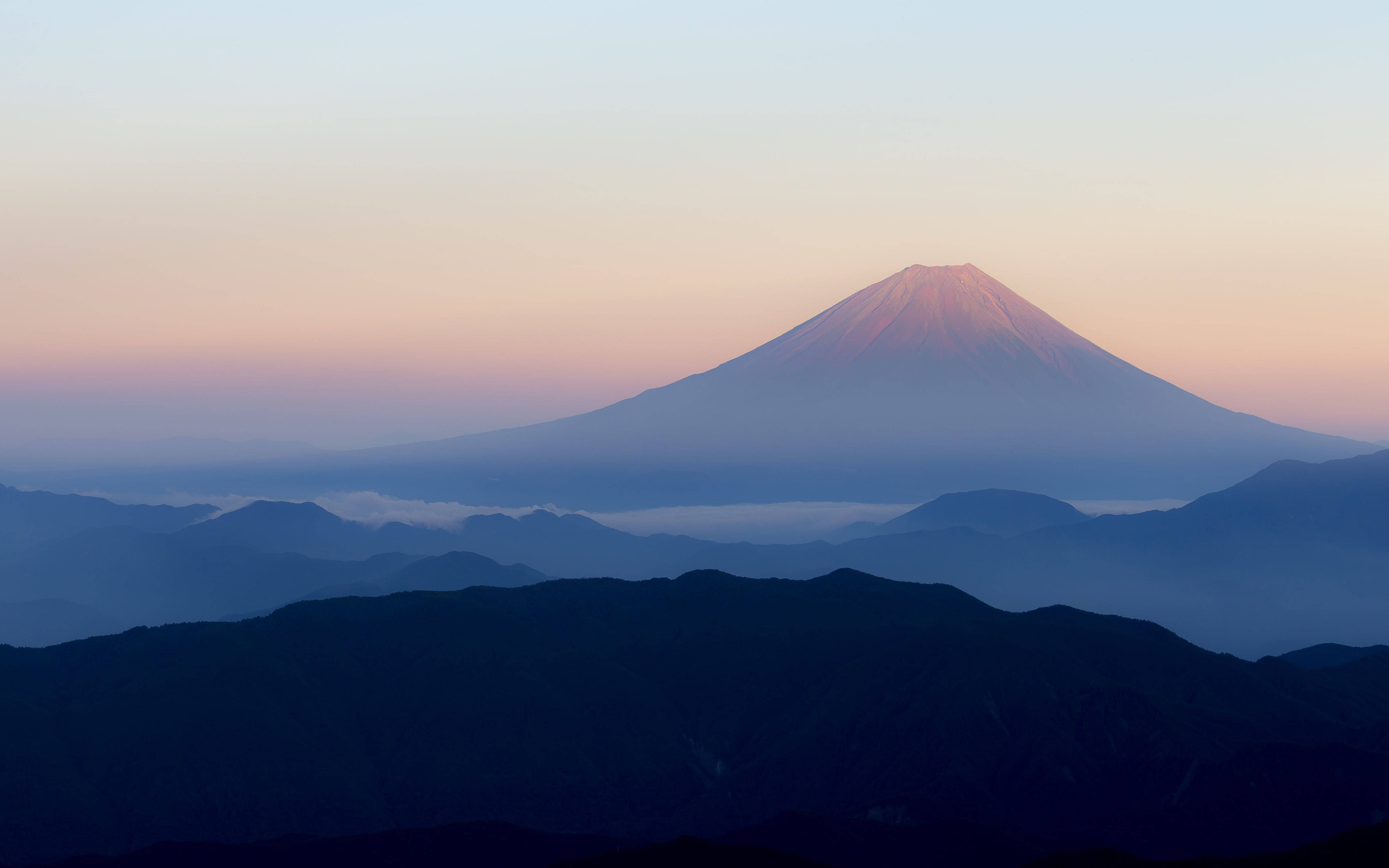 日本 桌布 壁紙 — 富士山在黎明時分雄偉地升起，柔和的粉紅色和藍色色調在層疊的山巒輪廓上空繪出天空。