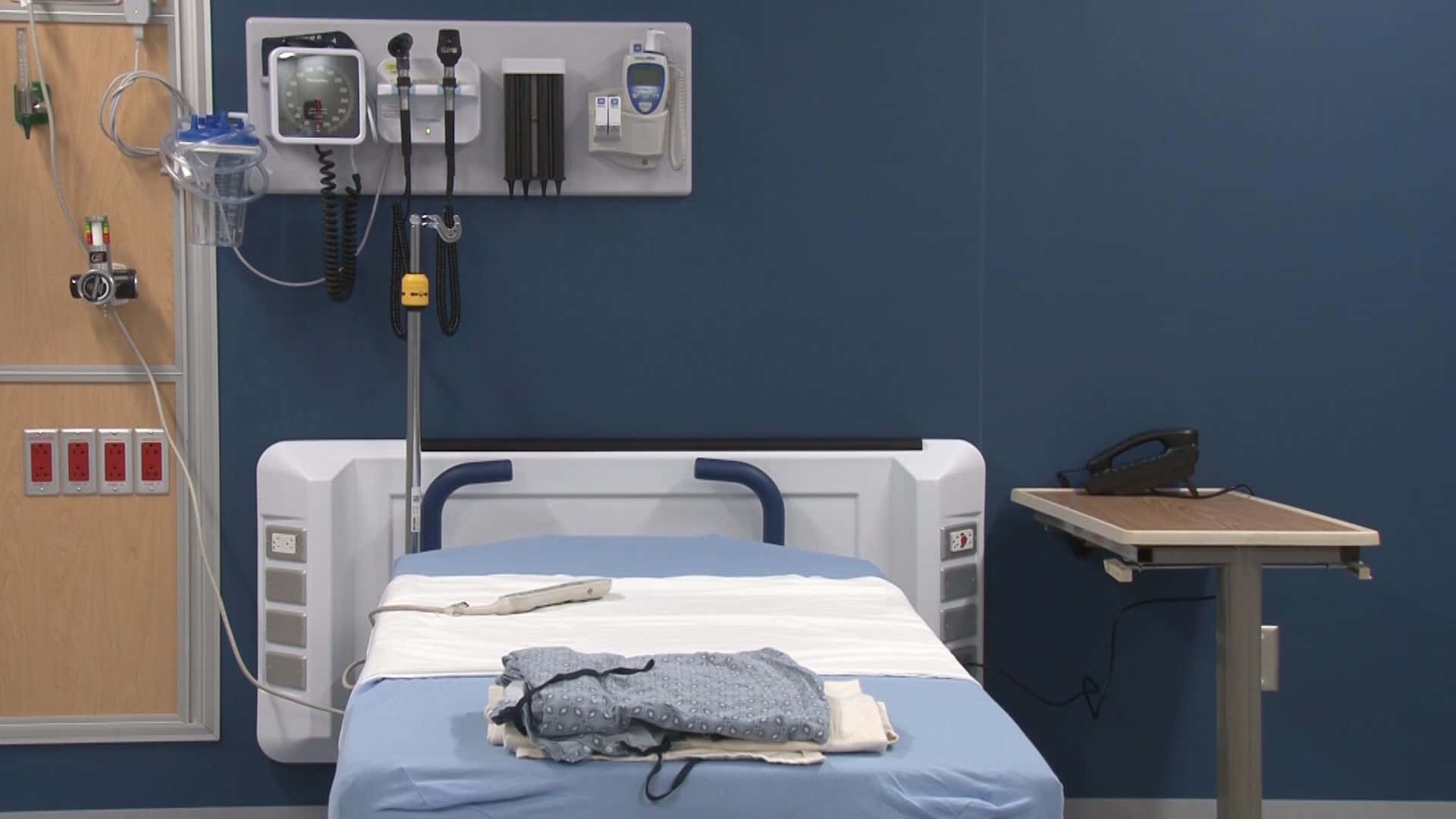 Hospital 桌布 壁紙 — 寧靜房間內的醫院病床，藍色牆壁上安裝了醫療設備，床上放有摺疊好的衣物。