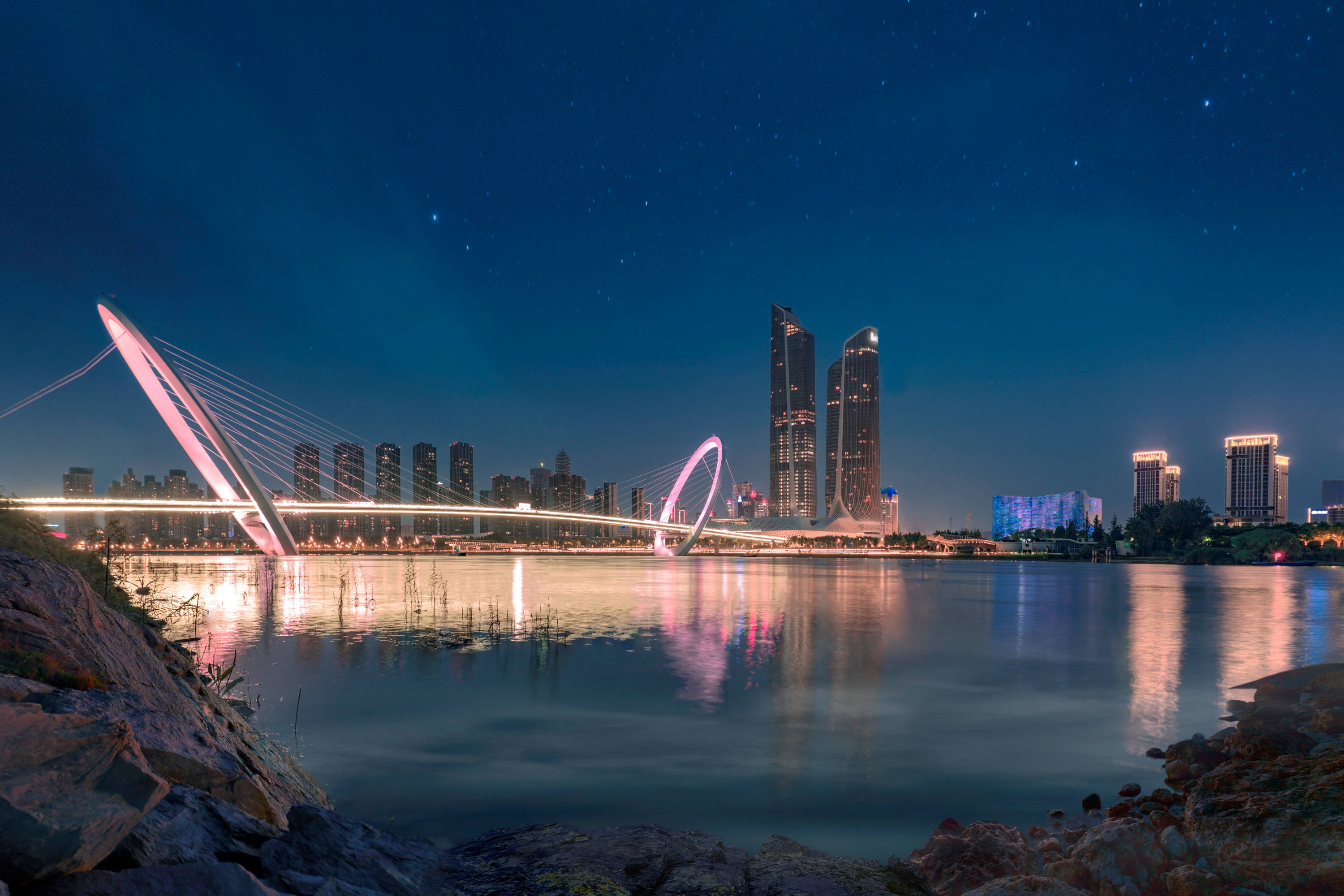 Nanjing 桌布 壁紙 — 寧靜水面上，一座發光的現代橋樑與城市摩天大樓及星空倒影的絕美夜景。