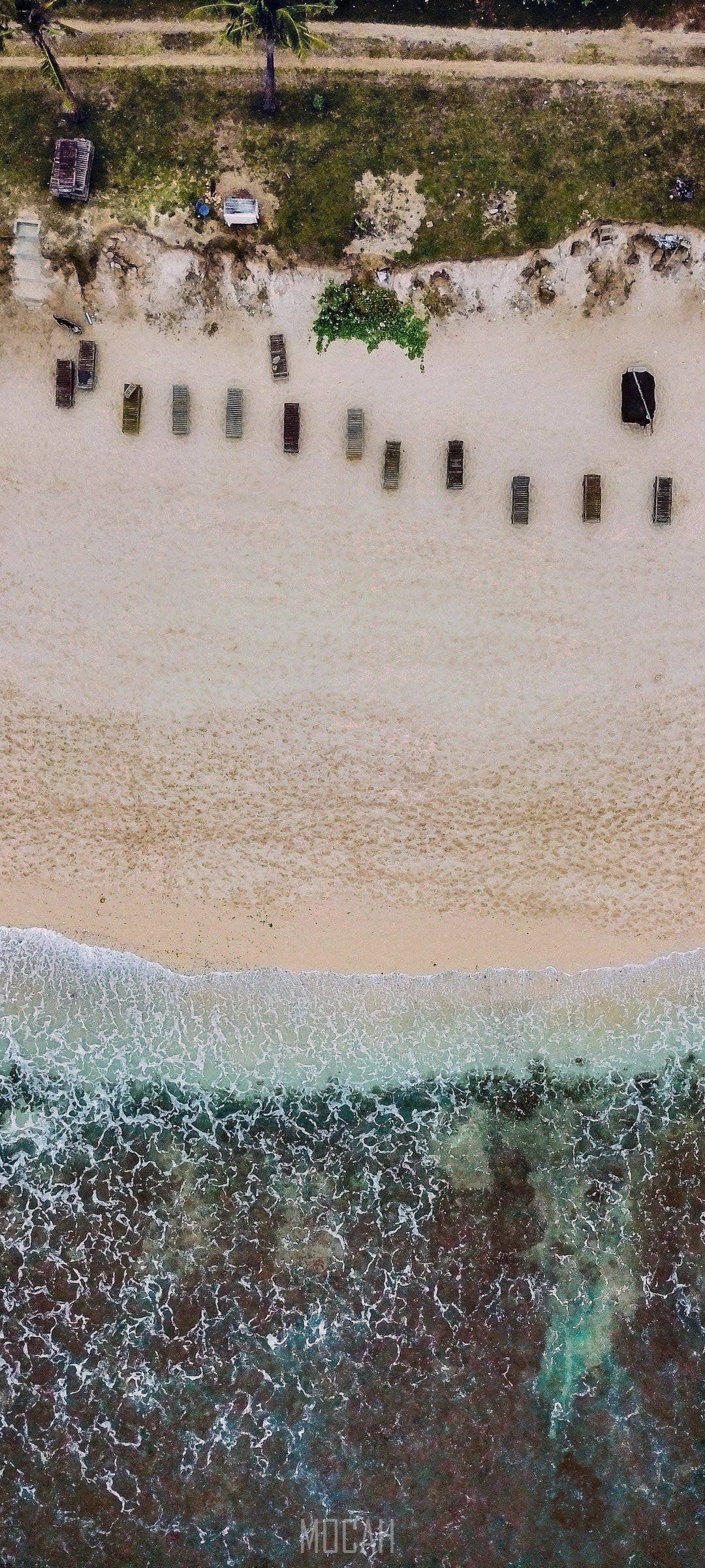 三星 桌布 壁紙 — 寧靜海灘的鳥瞰圖，一排排日光浴躺椅面向碧綠的海浪和長滿草的海岬，海岬上有棕櫚樹。