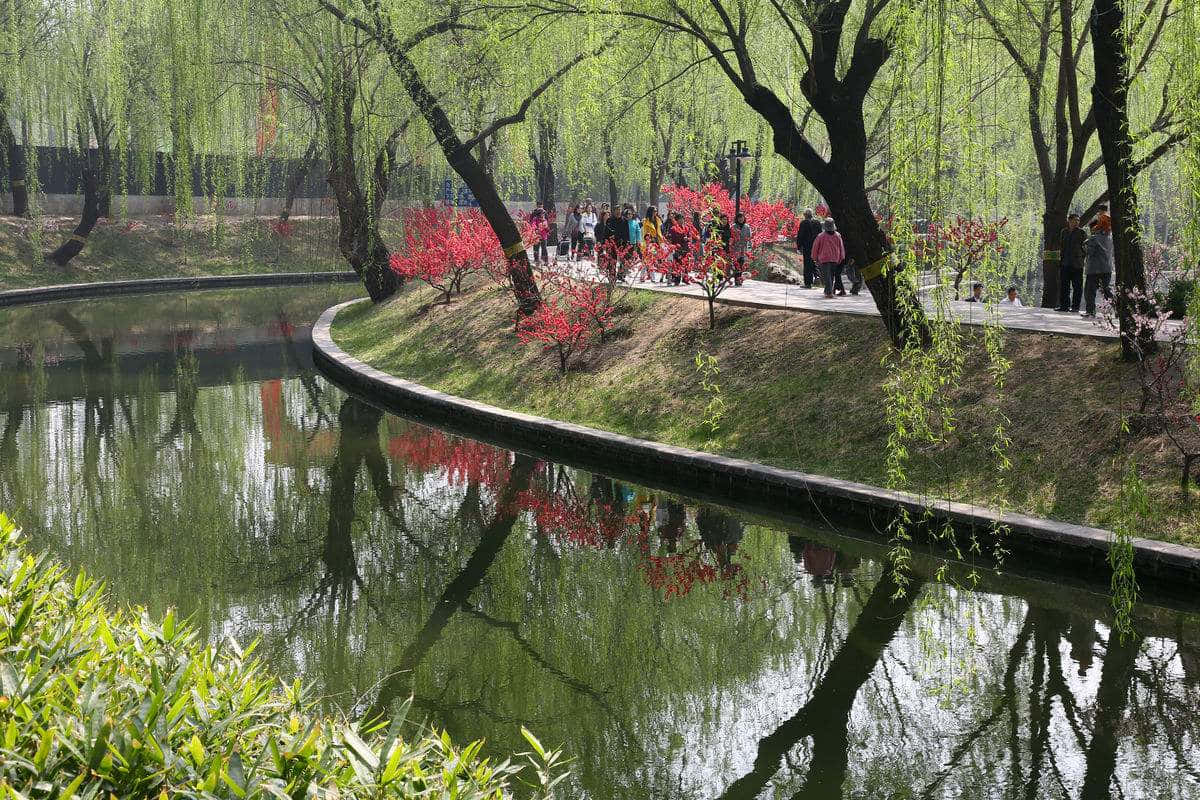 公園 桌布 壁紙 — 寧靜的公園場景，蜿蜒的運河倒映著垂柳和鮮紅色的開花樹木，人們沿著小徑漫步。