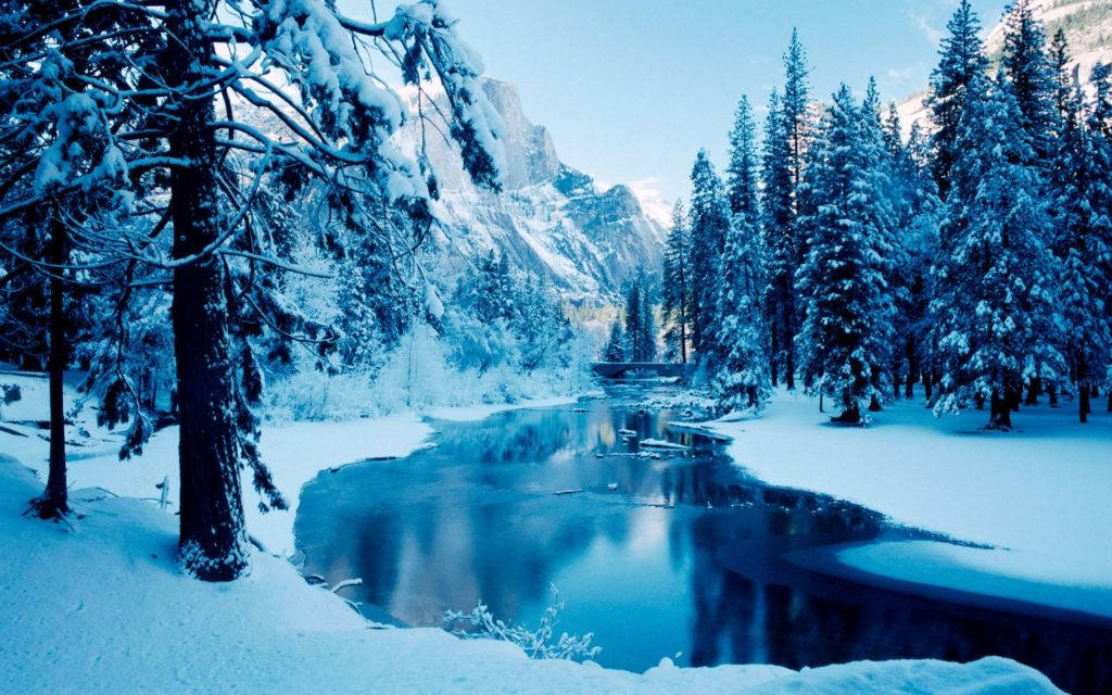 冬季桌面 桌布 壁紙 — 寧靜的冬日景象，白雪覆蓋的松樹排列在平靜的河流兩旁，河流映照著冰藍色的山脈，天空清澈。