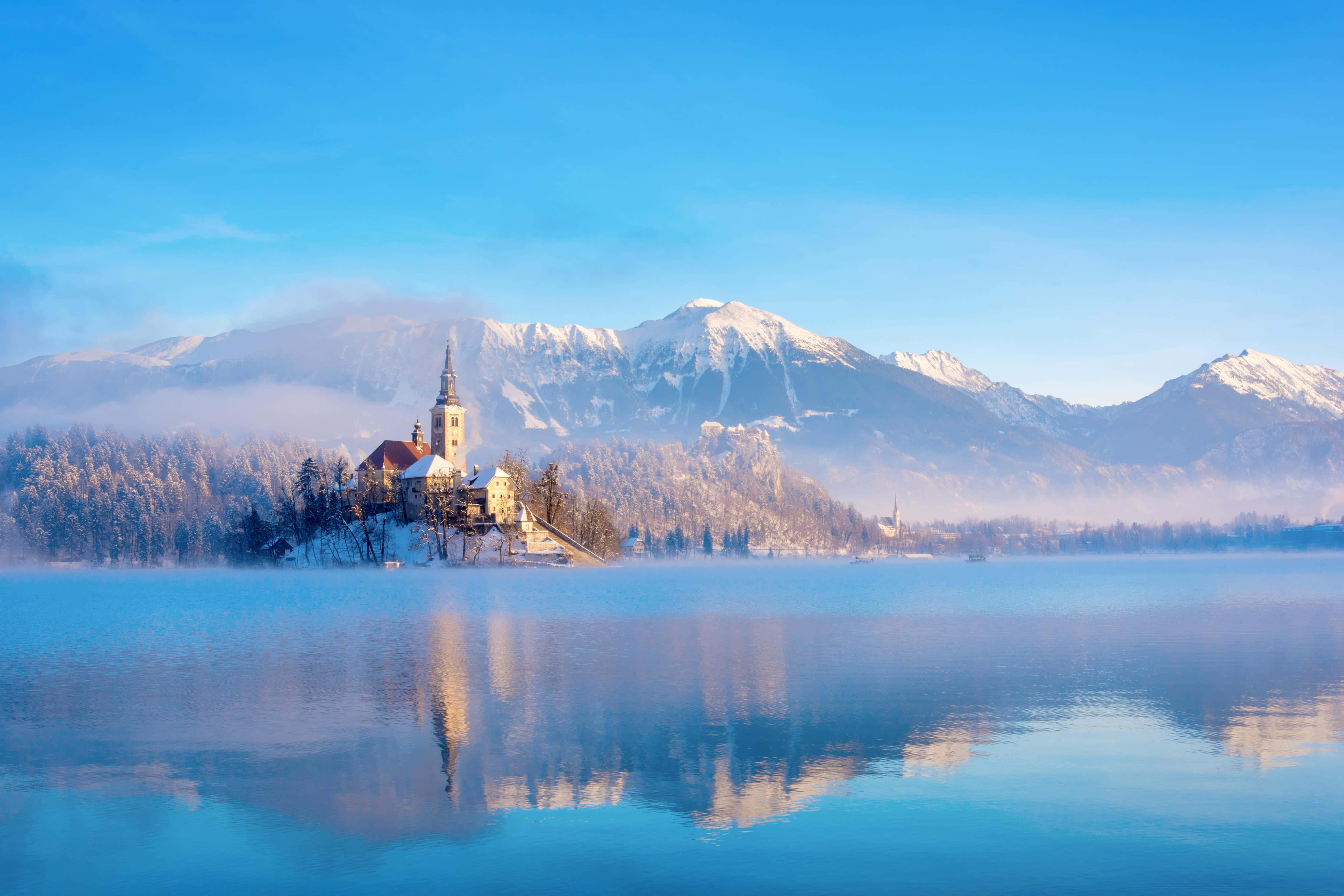 Alps 桌布 壁紙 — 寧靜的冬日湖泊映照出島上覆蓋著雪花的教堂，背景是霧氣繚繞的山脈和清澈的藍天。