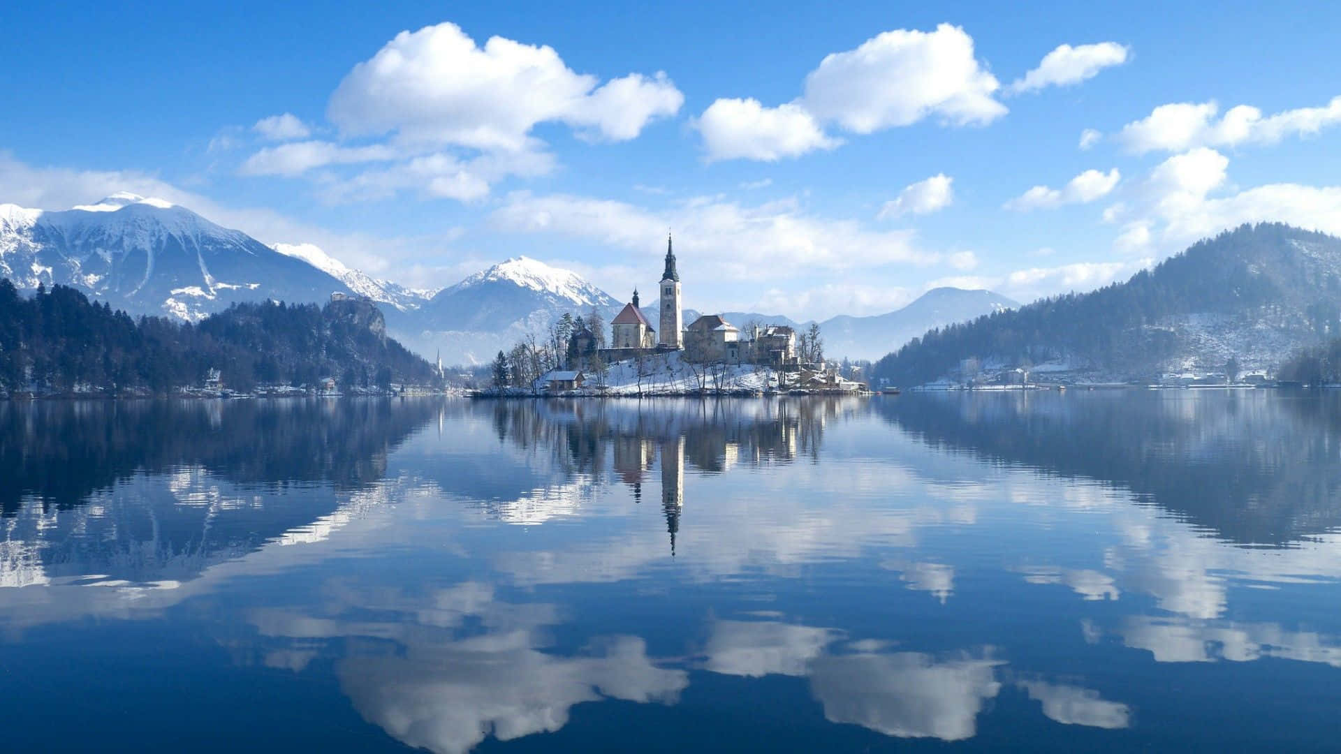 Lake Bled 桌布 壁紙 — 寧靜的冬日湖泊映照著白雪覆蓋的山脈和一座小島上的歷史教堂，天空湛藍。
